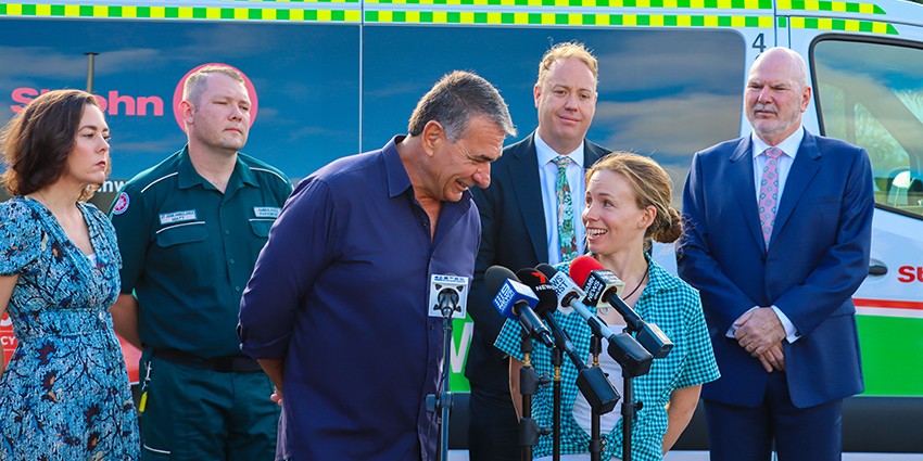 Image of Cassie Rowe MLA, Matt Didcoe, Rod Balkovitch, Nicole Tolhurst, Minister David Michael, Adrian Warner standing in front of microphones with an ambulance in the background