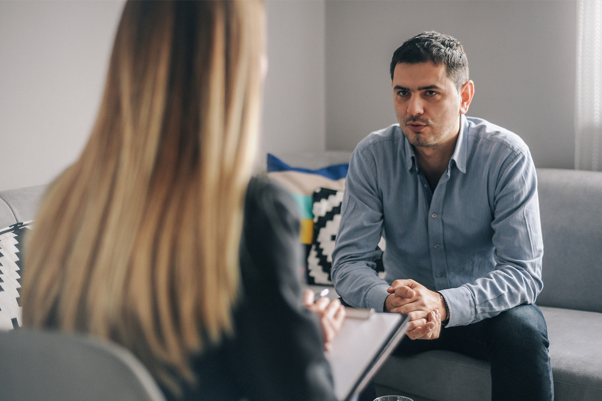 Man sitting on a couch having a conversation with a woman also seated holding a clipboard.