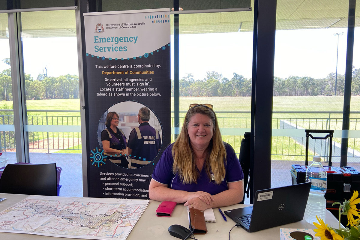 Member of the State Welfare Incident Coordination Centre at a help desk. There is a map and laptop on the desk, with an Emergency Services information banner behind the desk.