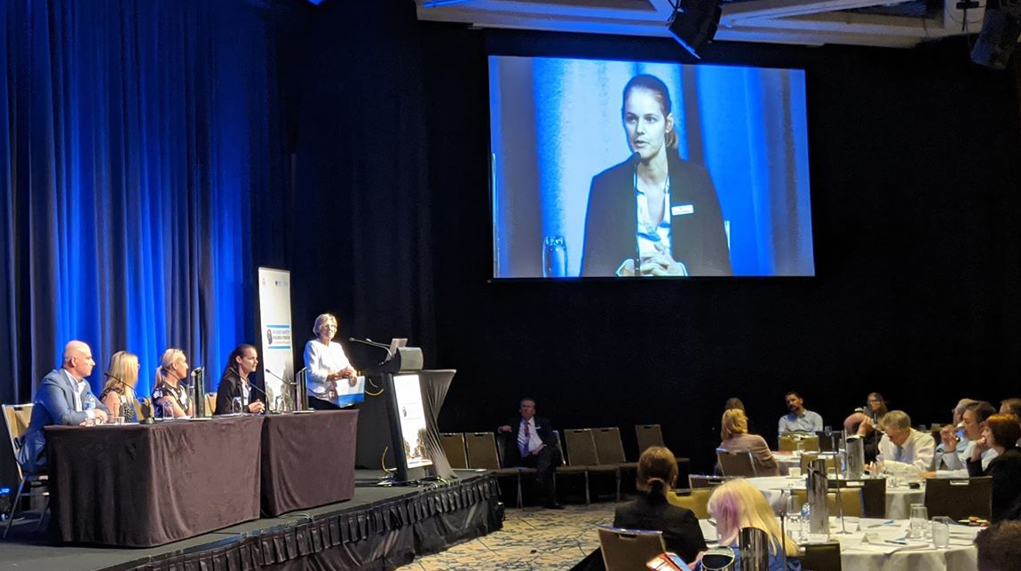 People speaking in a large room watching a speaker on a screen