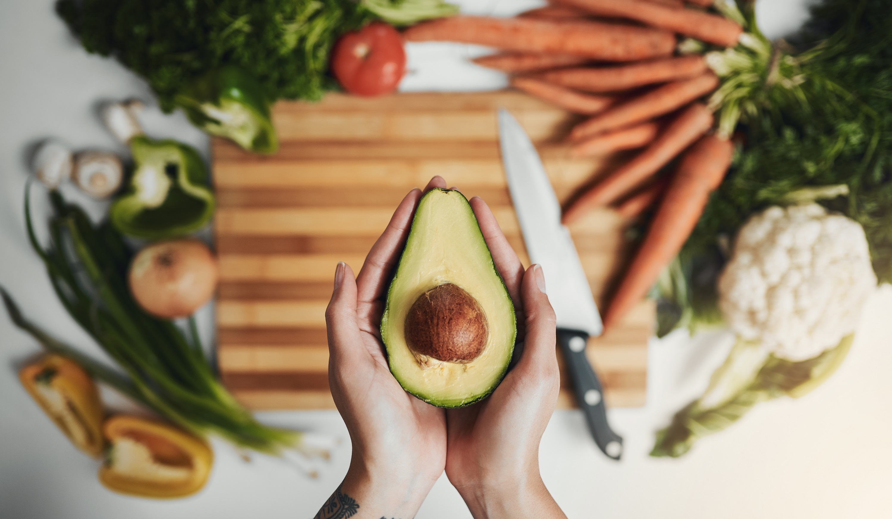 a womans hands holding a half avocado over a chopping board  with vegetables scattered around