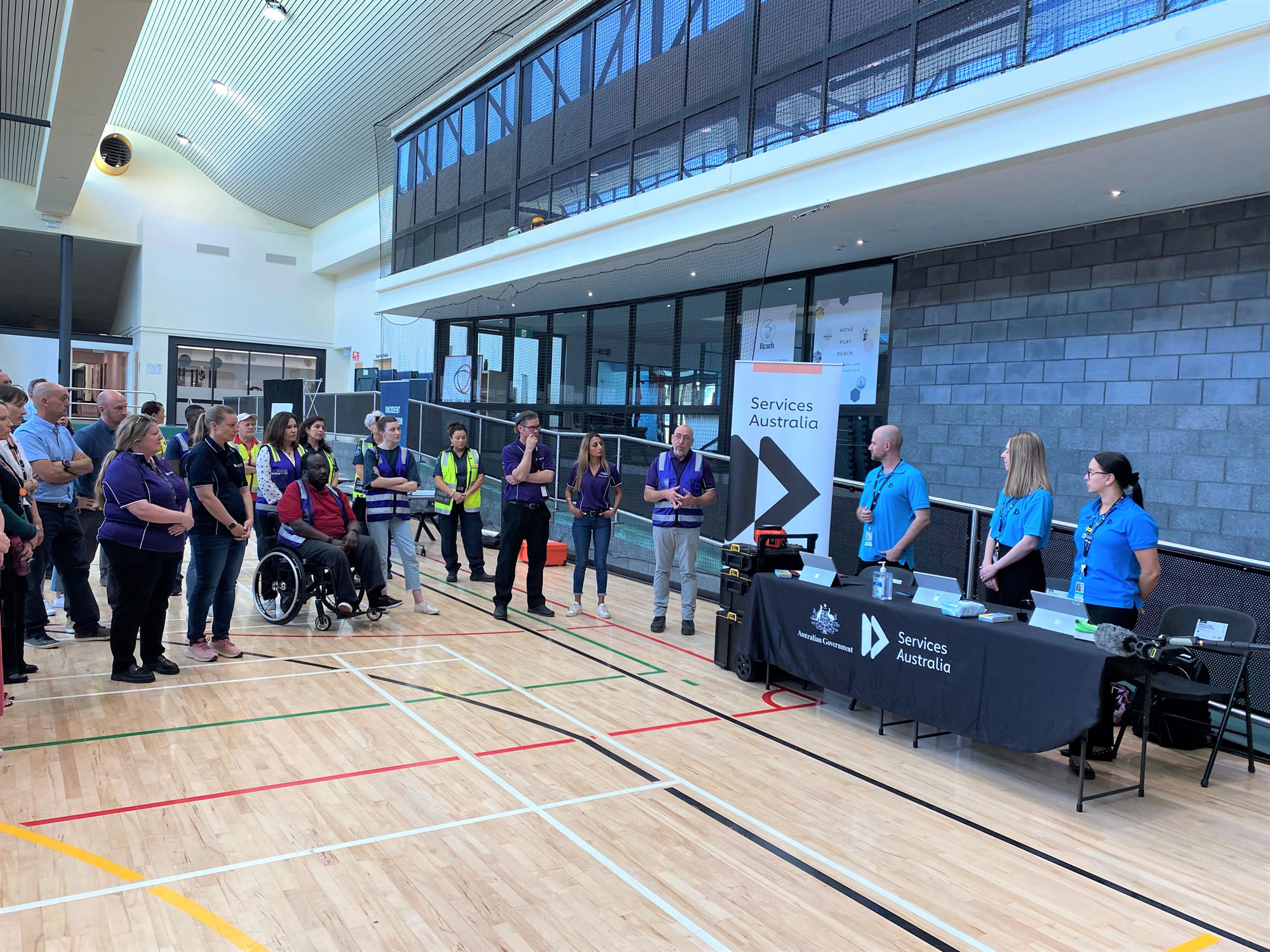 Group of emergency response trainees listening to man presenting on indoor basketball court. Signage in the photograph displays the words 'Services Australia'.