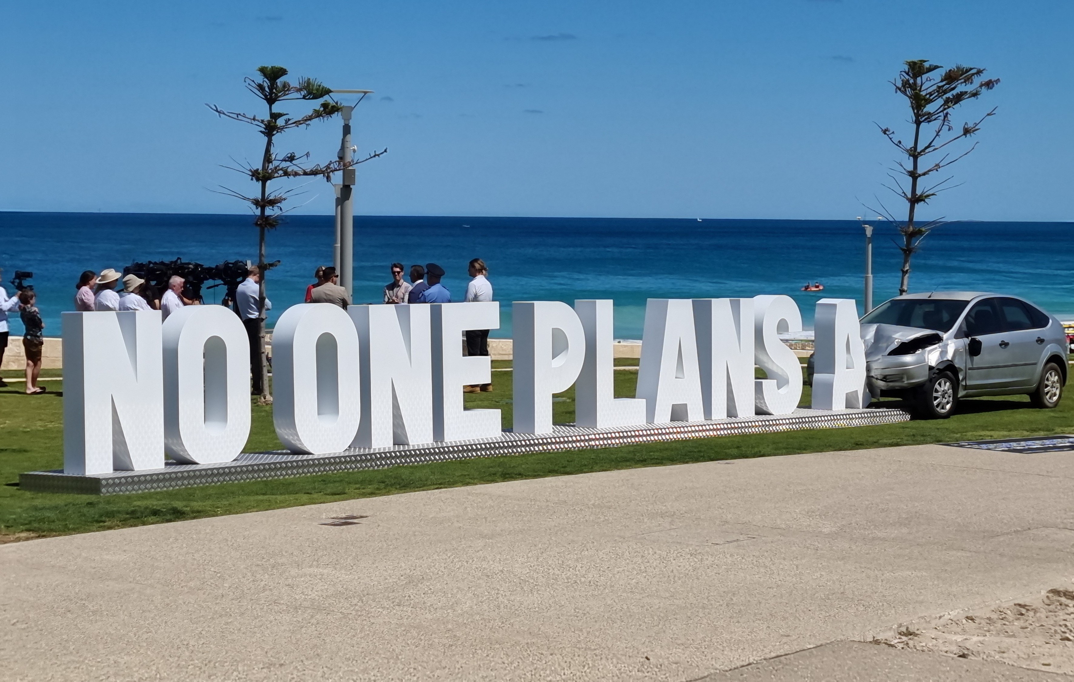 3d text reading 'no one plans a' next to a crashed vehicle, as an art installation with a beach in the background