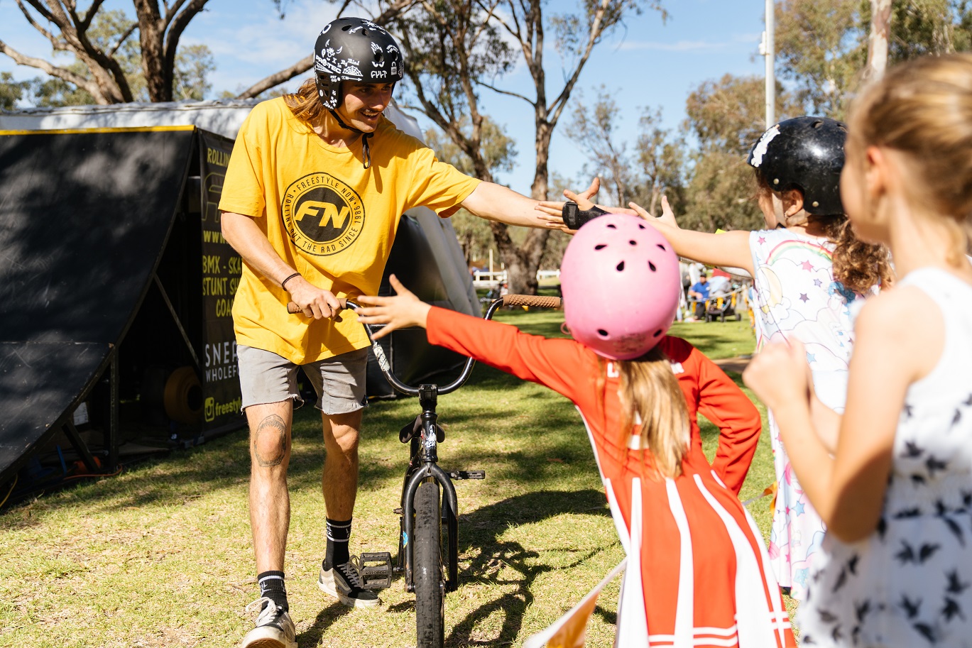 a man wearing a yellow shirt wheels a bicycle on some grass, as he passes a group of children he puts his arm and hand out for a high five.
