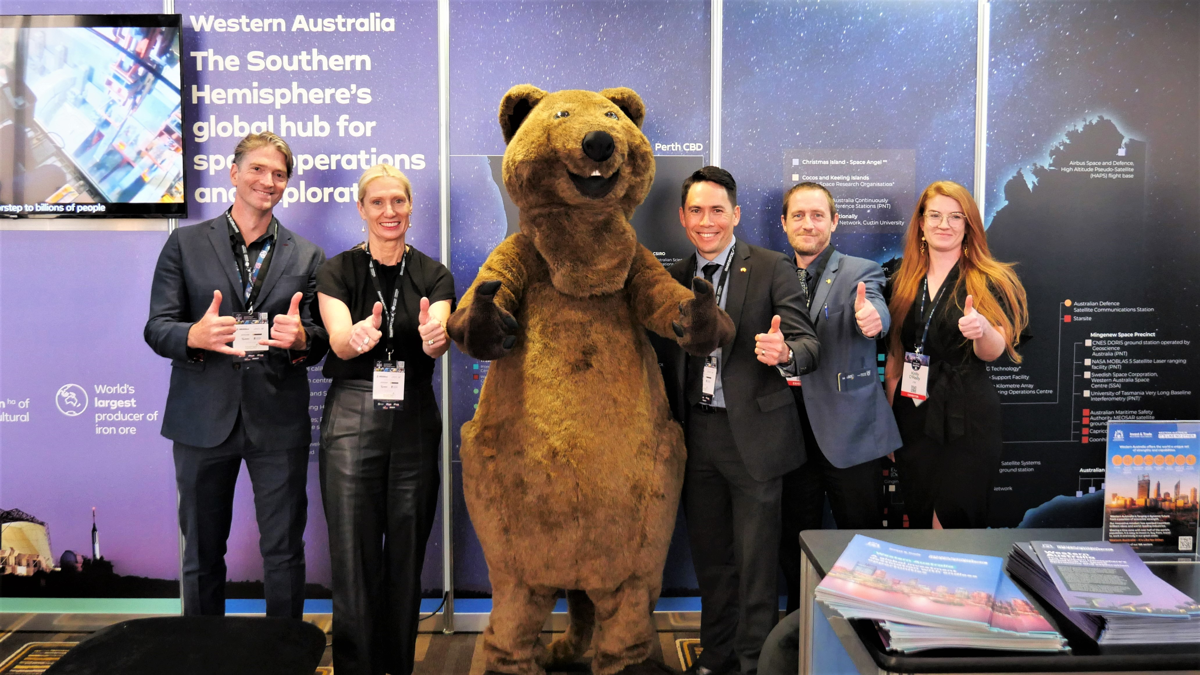 A group photo in front of the Department of Jobs, Tourism, Science and Innovations booth. From left to right is Ash Boddy, Linda Dawson, A person in a Quokka suite, James Yuen, Simon Aaron and Kirilly O'Reilly. Everyone has their thumbs up.