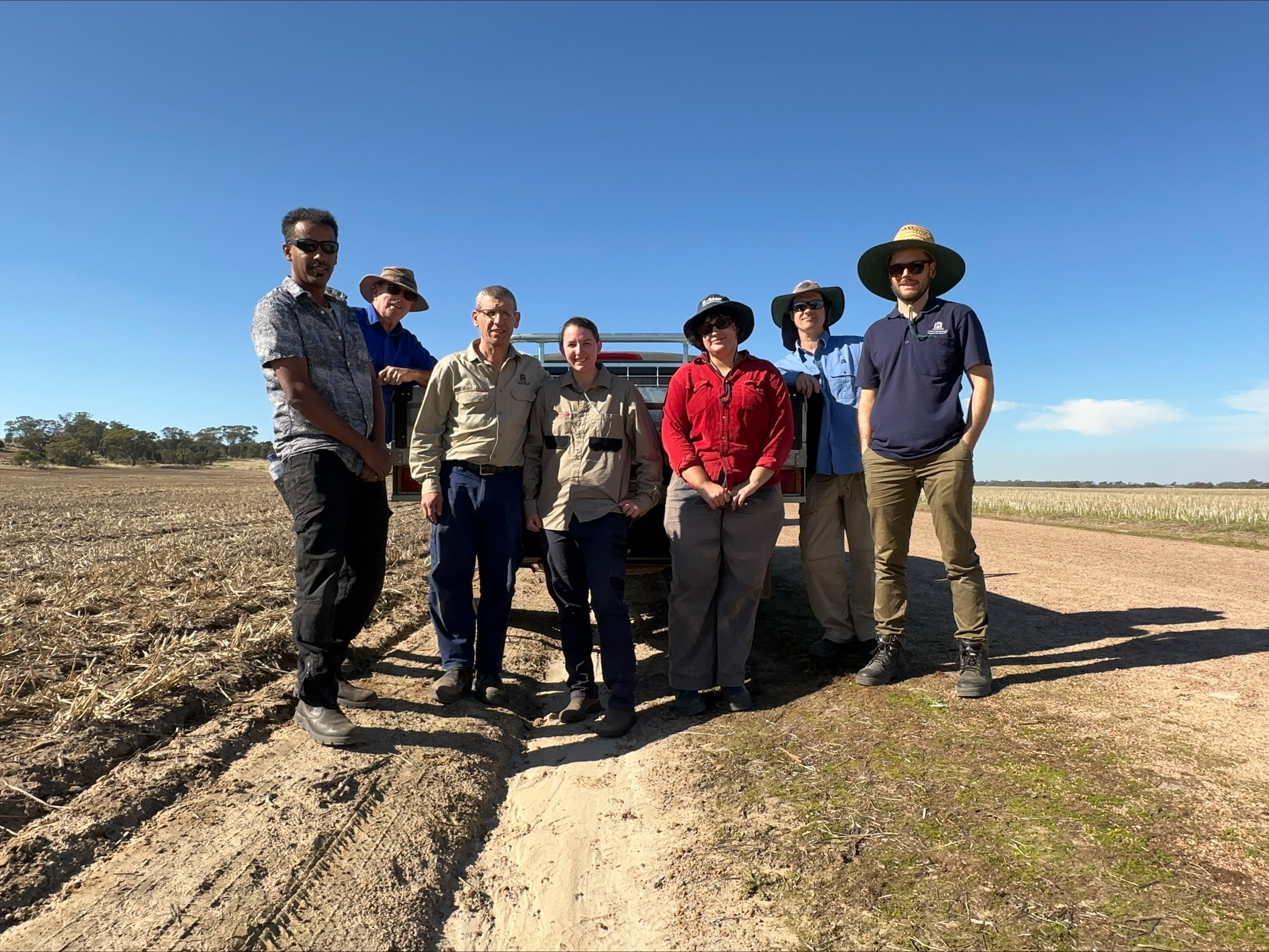 A group of seven people standing in front of a ute in a dry paddock.