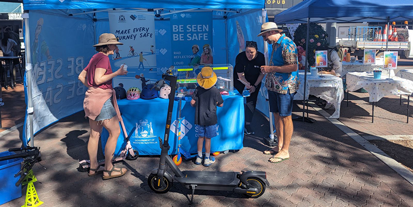 Commission staff answering questions at a display on eRideables, a woman stands by a table and banner that says 'Be Seen Be Safe' while talking to a couple with a child. An eRideable device stands in the foreground.