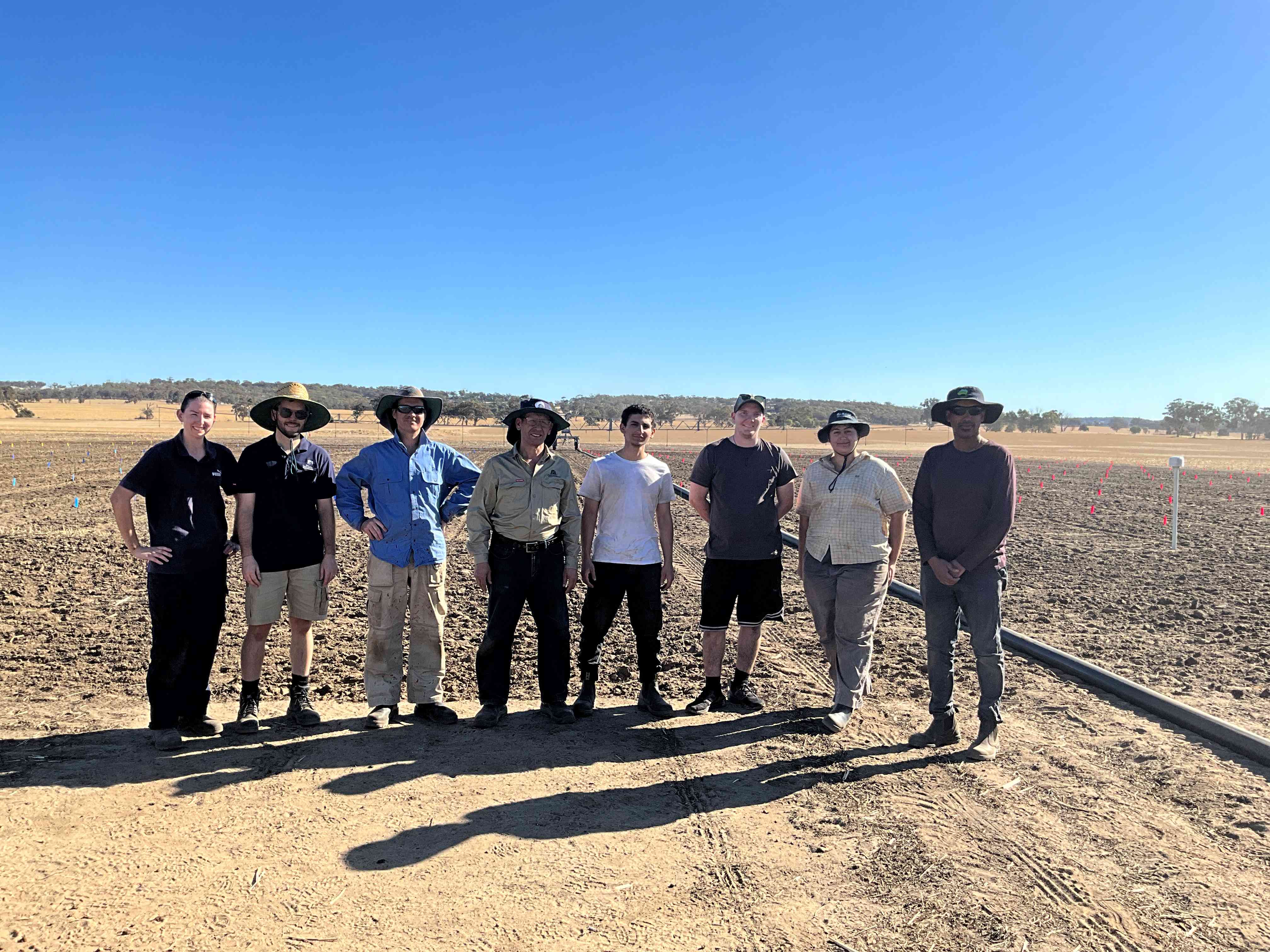 Eight people standing in a row in a dry paddock.