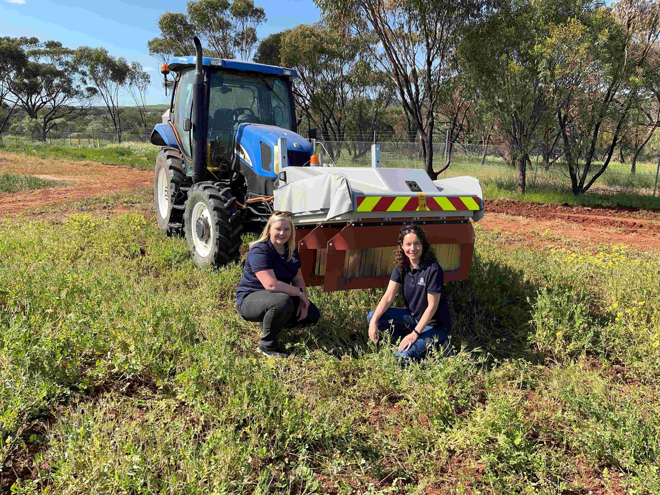 Two women crouching in front of a tractor with an implement. 