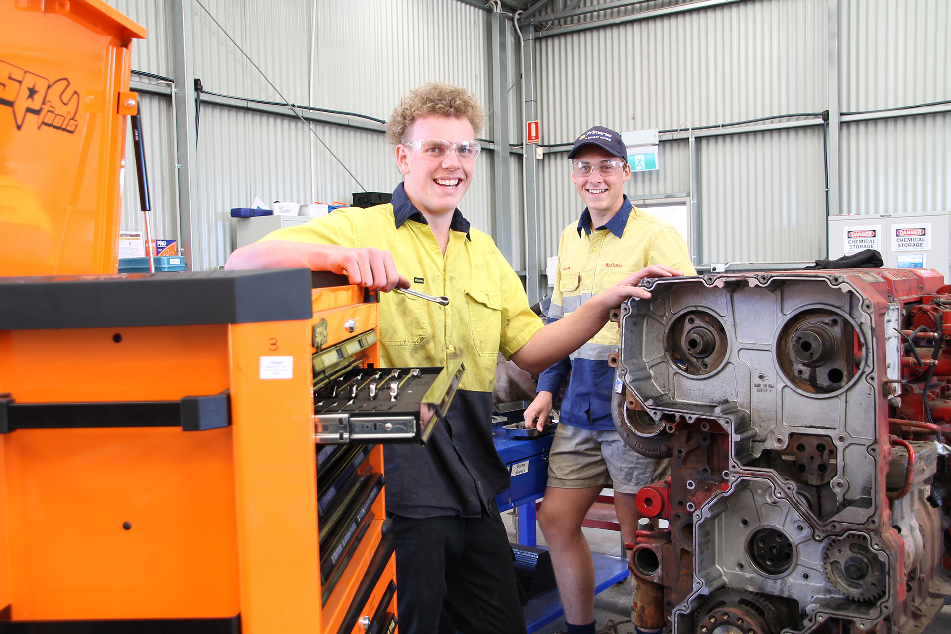 Two mechanics in a workshop, smiling while working on an engine with a tool chest nearby.