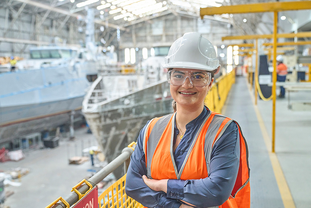 An engineer wearing high visibility workwear, stands in front of a marine vessel in a large workshop. 