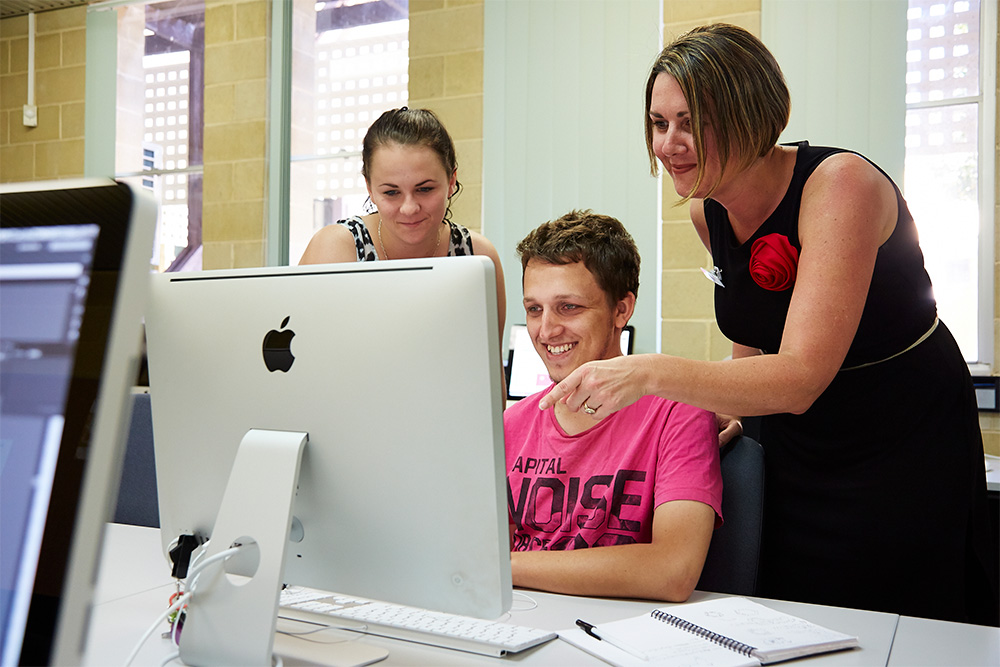 A lecturer supporting two students as they look at a computer screen.