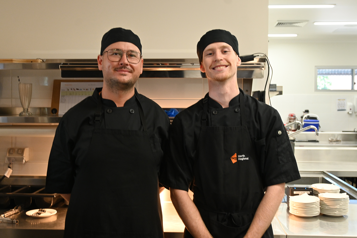 Two men in black aprons stand together in a kitchen, engaged in a cooking activity.
