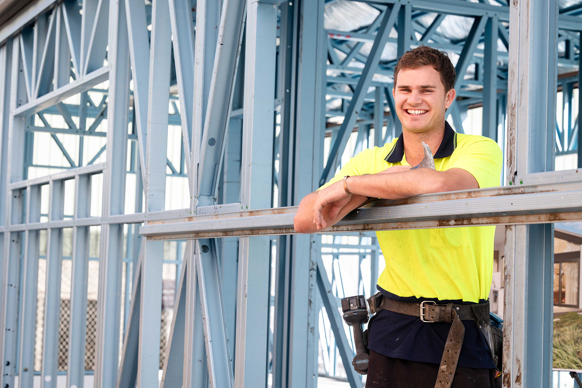 A man in a yellow shirt and safety vest stands in front of a steel frame, overseeing construction work.