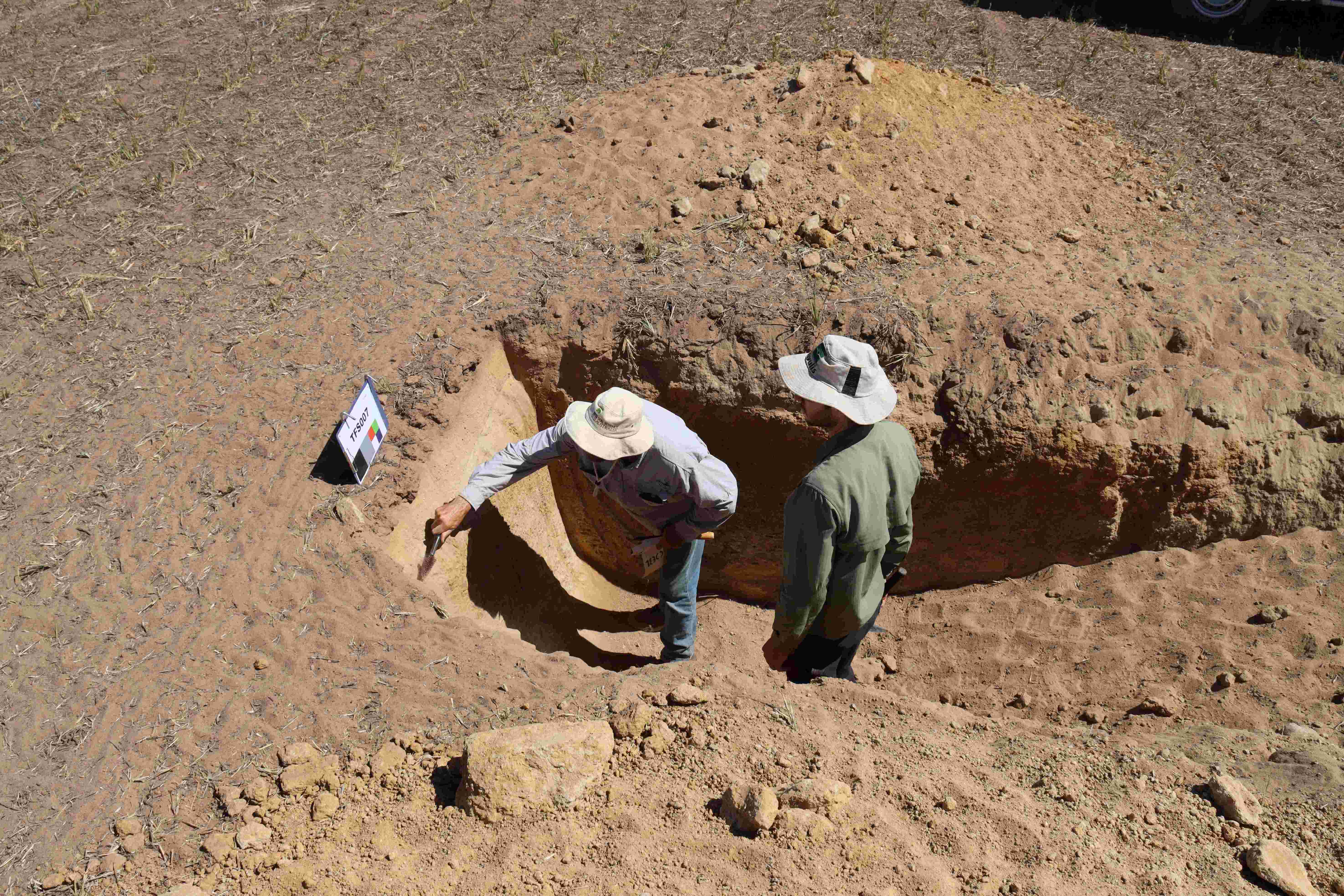 Two men in a hole looking at sand in a dry paddock.