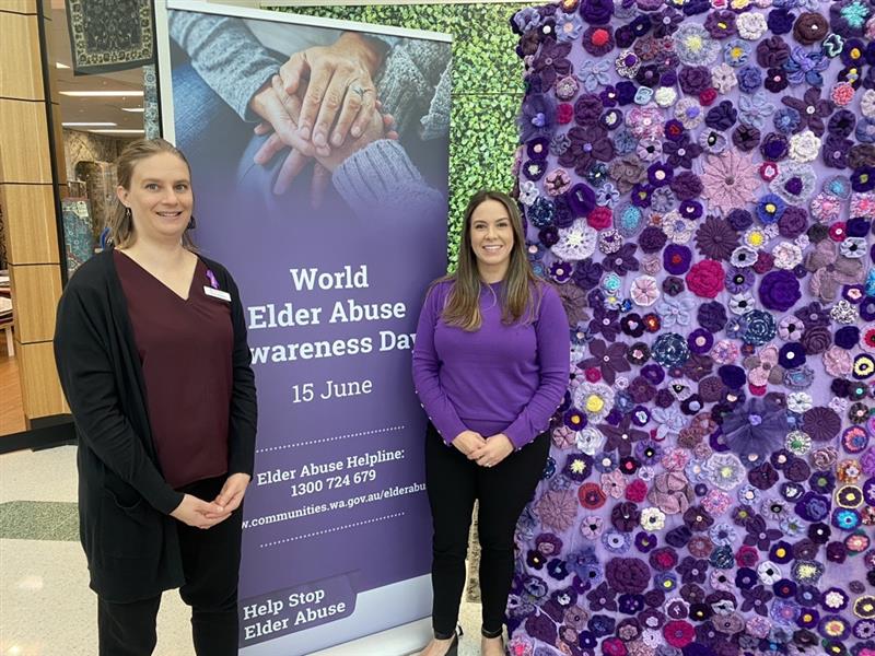 Image off two Communities staff members (two young women) standing next to the World Elder Abuse Awareness Day banner and Purple Road material