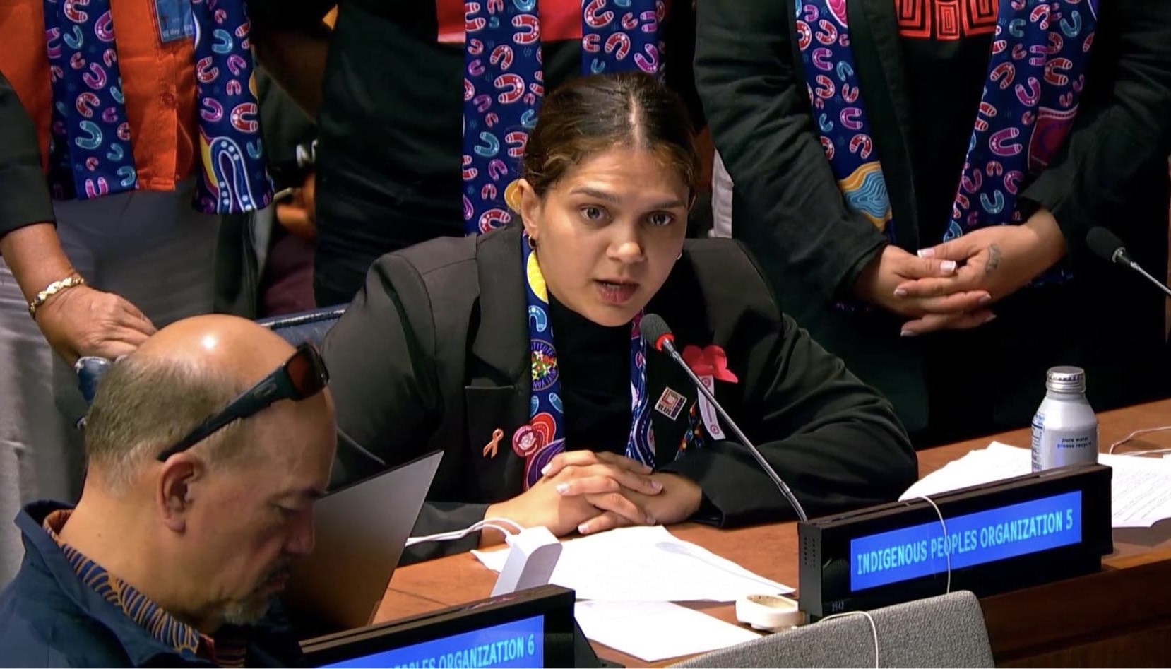 A young Aboriginal woman speaking into a microphone, with an audience watching on