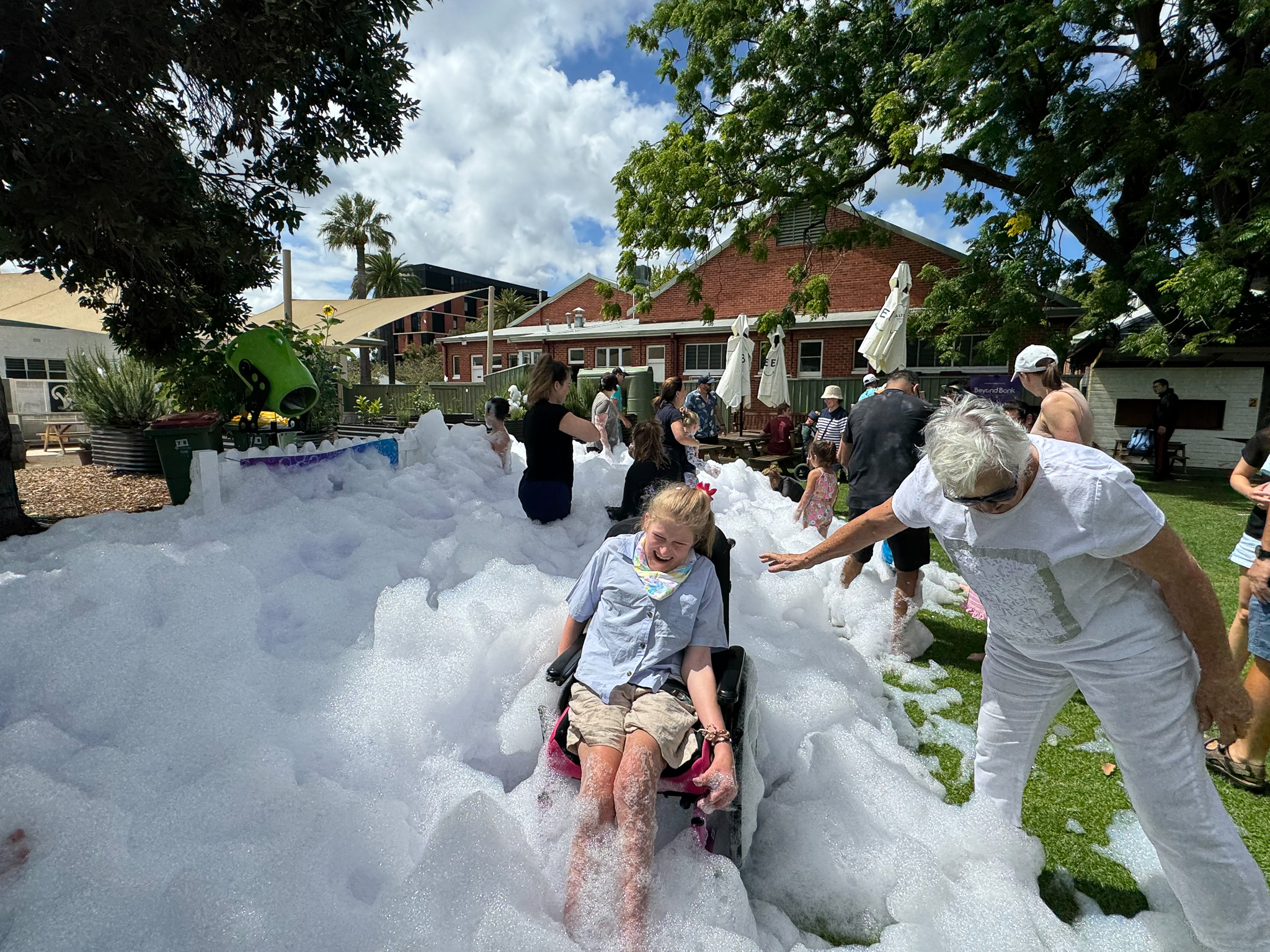 A lively outdoor foam party with children and adults playing in thick white foam spread across a grassy park. Some people are standing, others are sitting or lying in the foam. Trees and buildings are visible in the background under a partly cloudy sky.