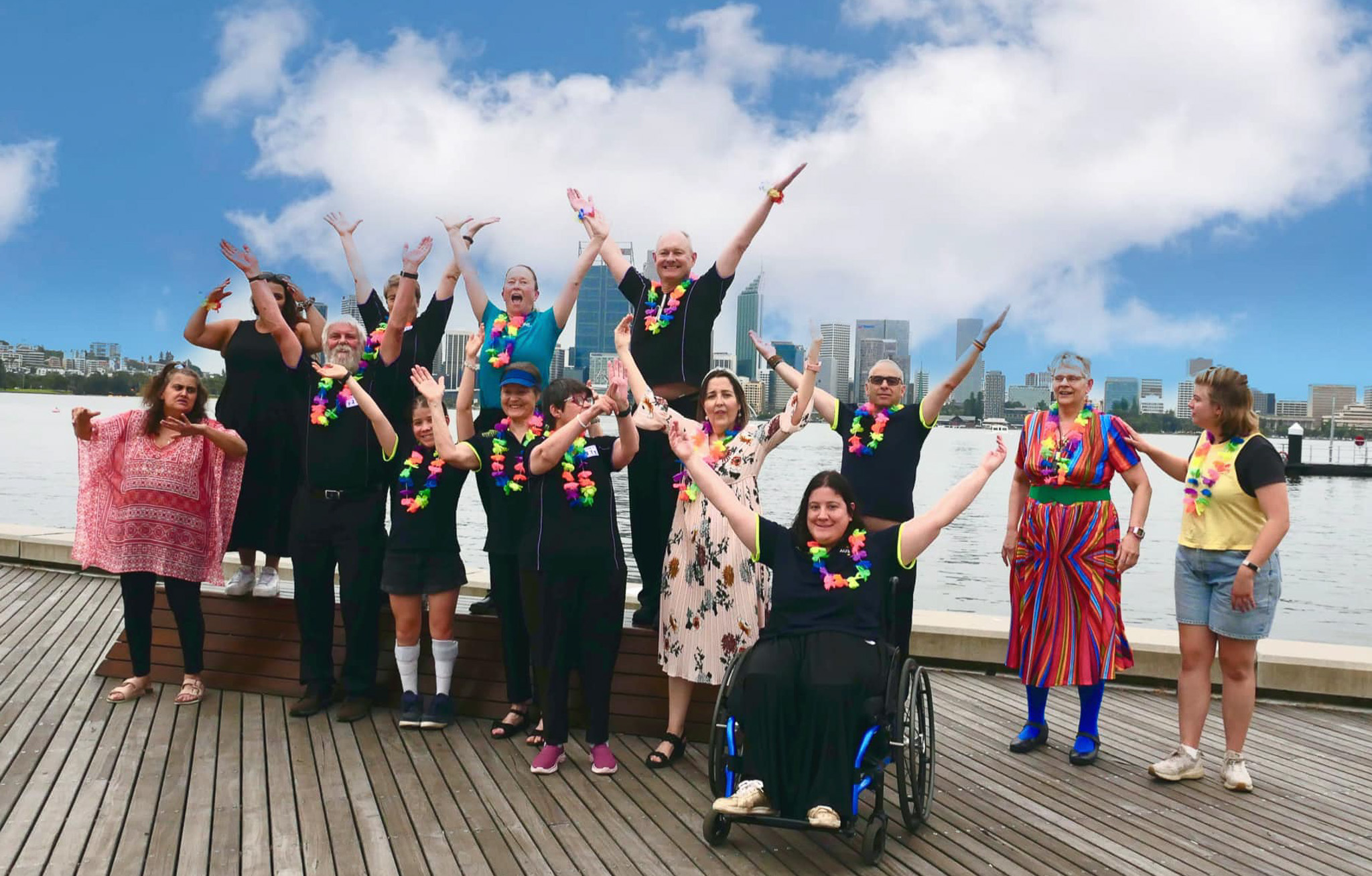 A cheerful group of people, including one person in a wheelchair, pose with raised arms on a wooden deck beside a waterfront. They wear colourful leis, and a city skyline is visible in the distance beneath a blue sky with scattered clouds.