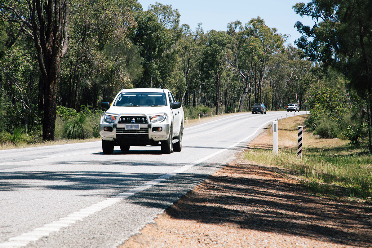 A white ute travelling down a carriageway with trees on either side. Two other vehicles are in the distance, travelling in the opposite direction.
