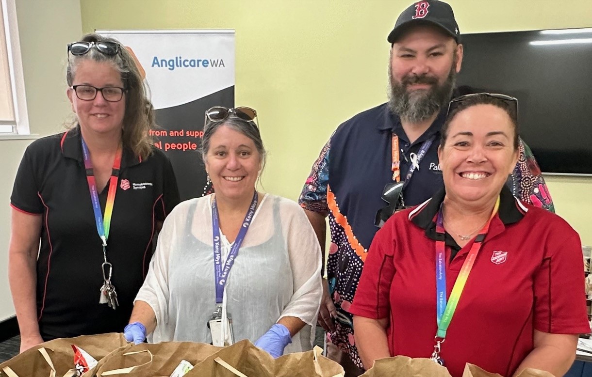 Four adults stand behind a table of brown paper bags filled with food, wearing uniforms and lanyards, with an Anglicare WA banner in the background.