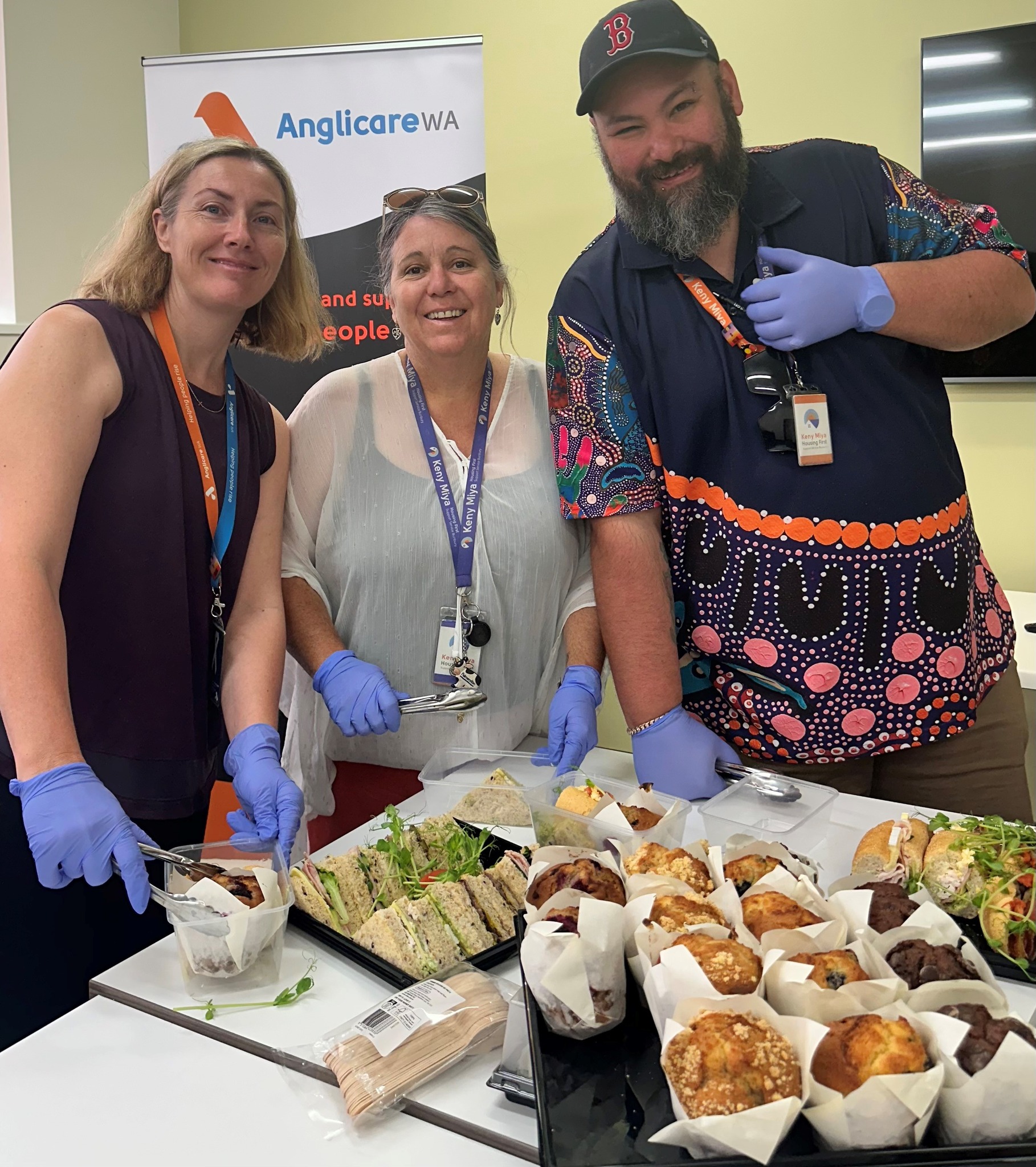Three smiling people in gloves stand behind a table of sandwiches, muffins, and baked goods, with an Anglicare WA banner in the background.