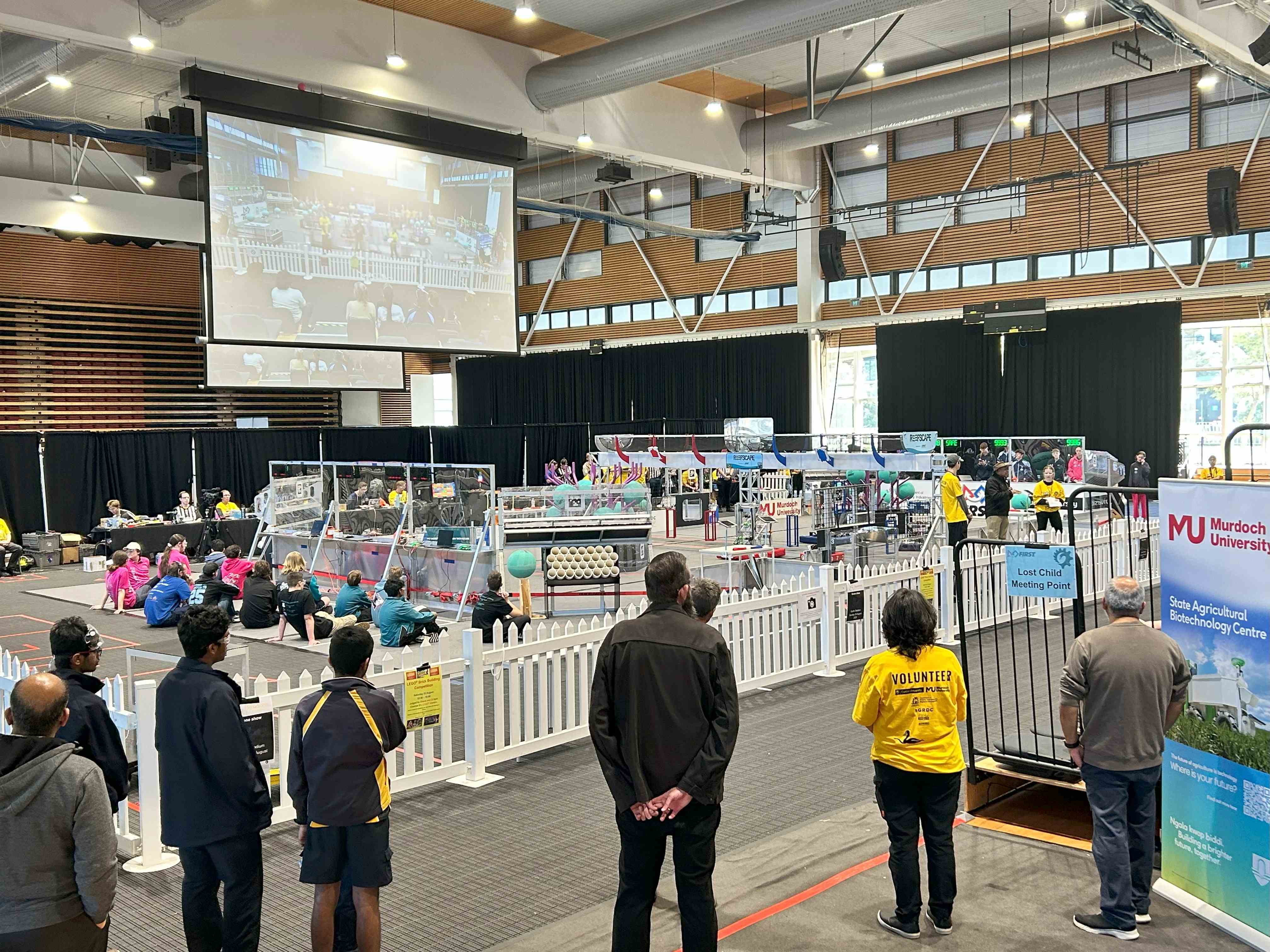 A fenced off area around robot equipment and children, shrouded by a big screen in front of an audience.