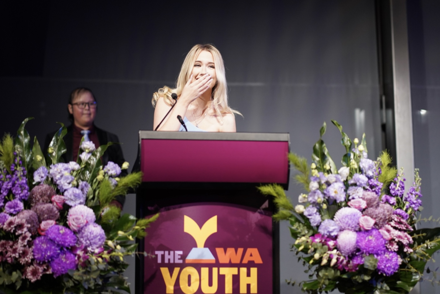 A young person at a podium with flowers, smiling and covering their mouth during the 2025 WA Youth Awards..