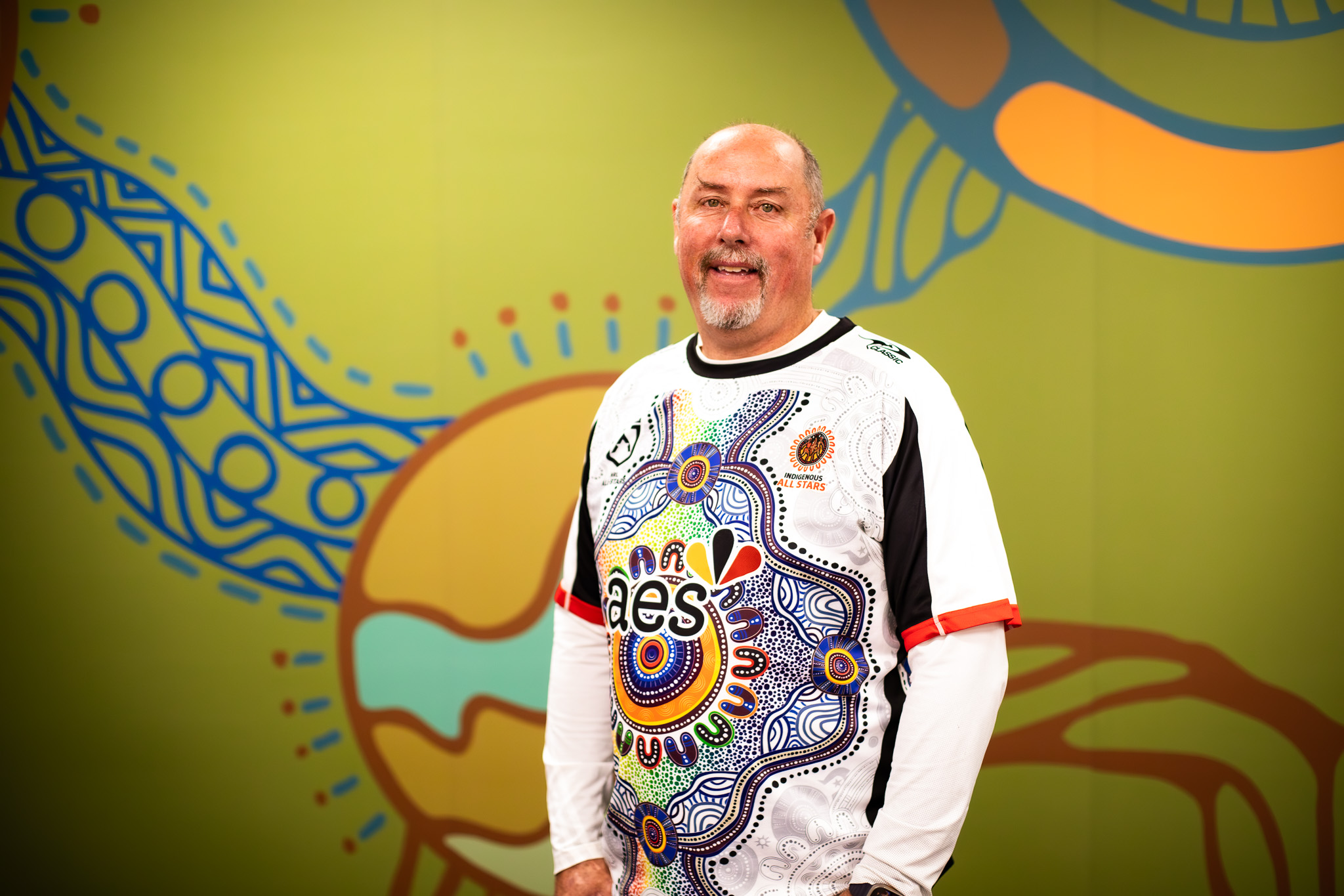 Image of a middle age man smiling and wearing a shirt with Aboriginal artwork