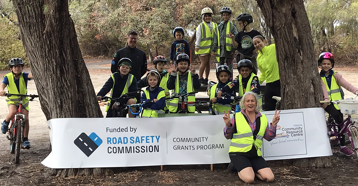 A group of children with bicycles amongst trees smiling and posing with a woman in the foreground in front of a banner that says "Funded by Road Safety Commission Community Grants Program"