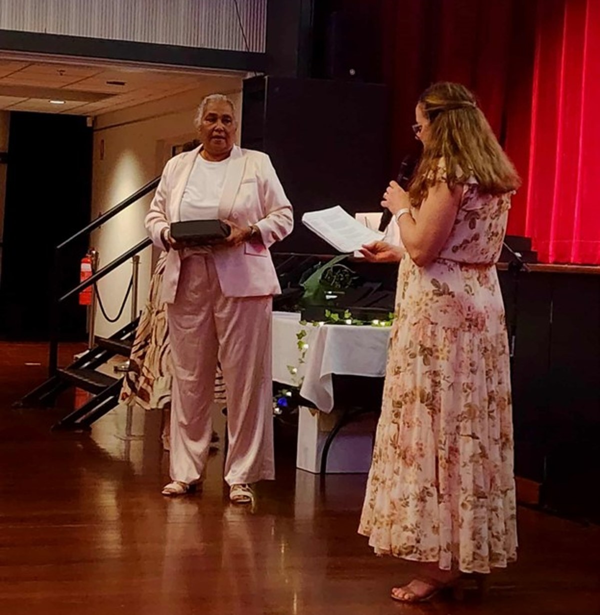 Two women are on a stage. One is receiving an award, the other is holding a microphone and reading a piece of paper.