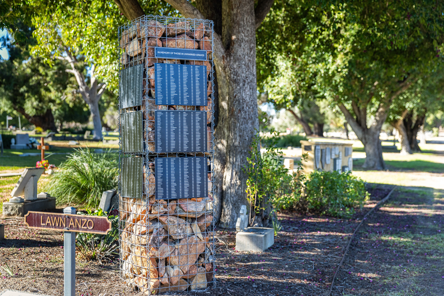 a gabion memorial pillar filled with rocks sits in a lush green garden. The pillar has a list of named of people buried in unmarked graves.