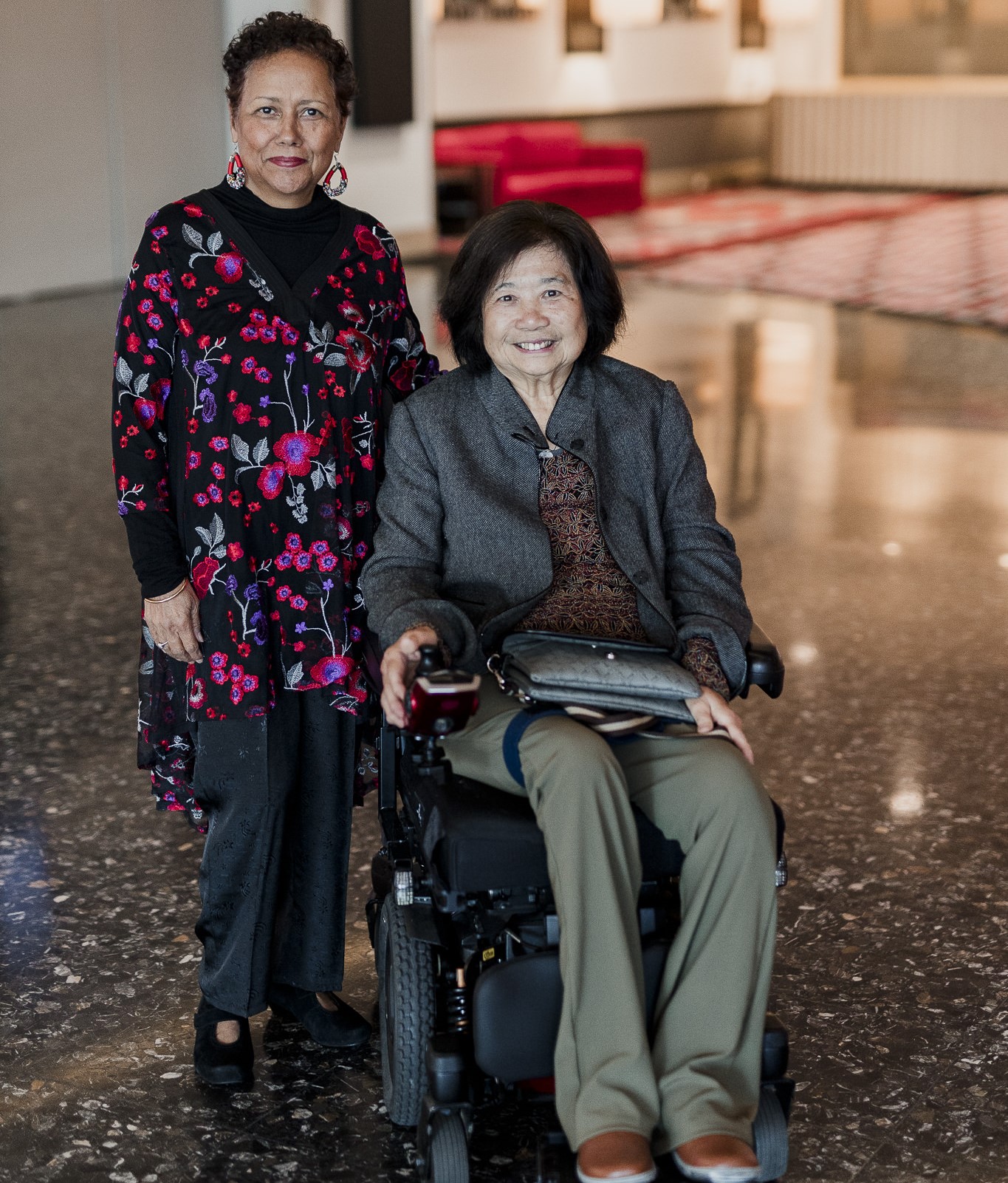 Two women, well dressed and smiling. One of the women is sitting in a wheelchair