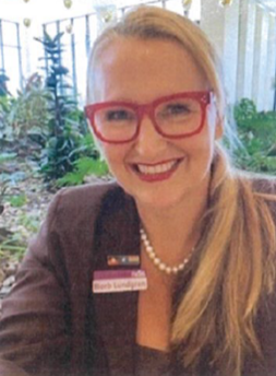 a well-dressed woman, smiling, wearing glasses, long blonde hair, pictured in an outdoor setting