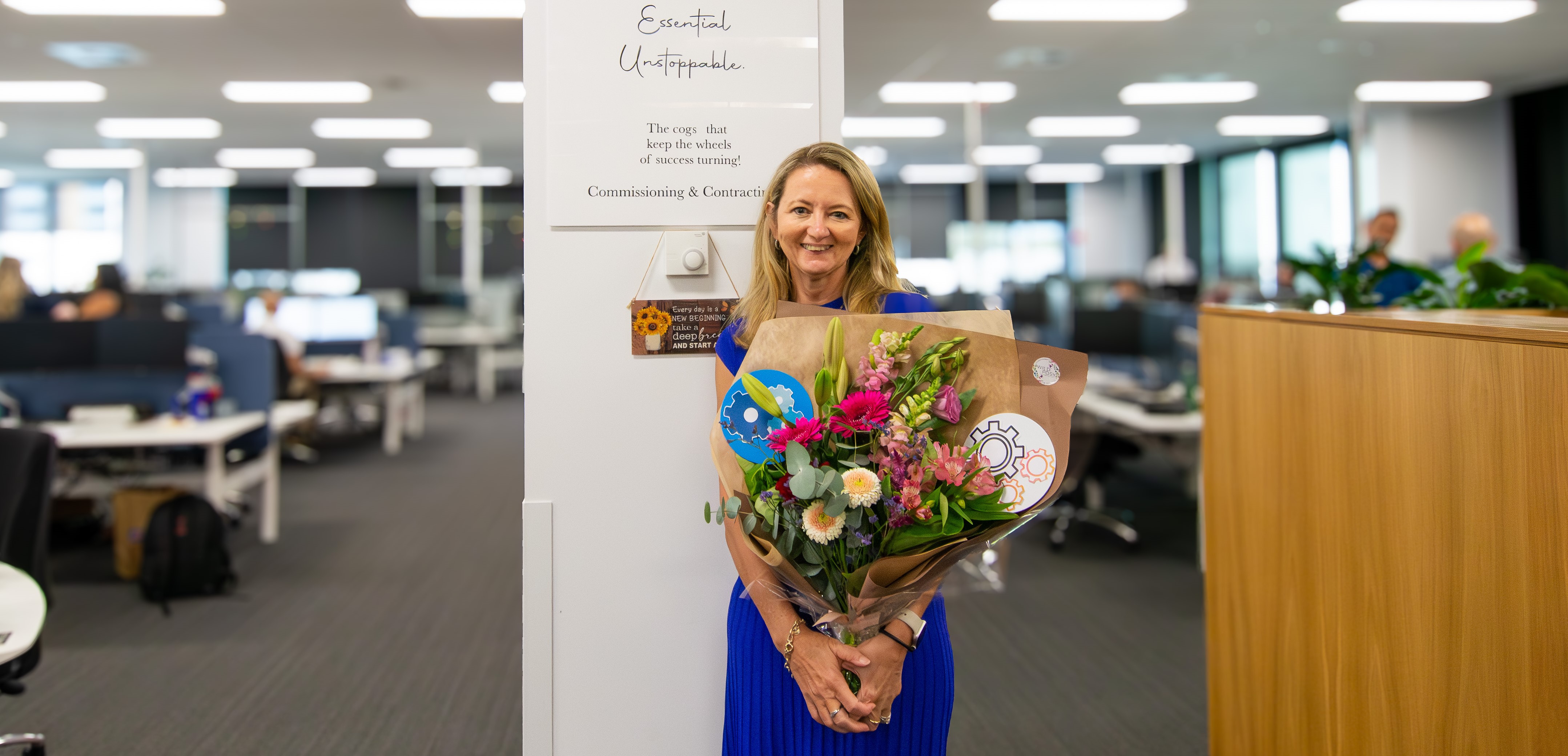 A woman in an office environment, smiling and holding a bunch of flowers