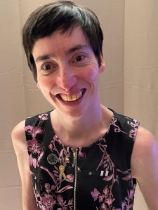 a woman, smiling, she has short dark hair, she's wearing a dress. Head and shoulders pictured.