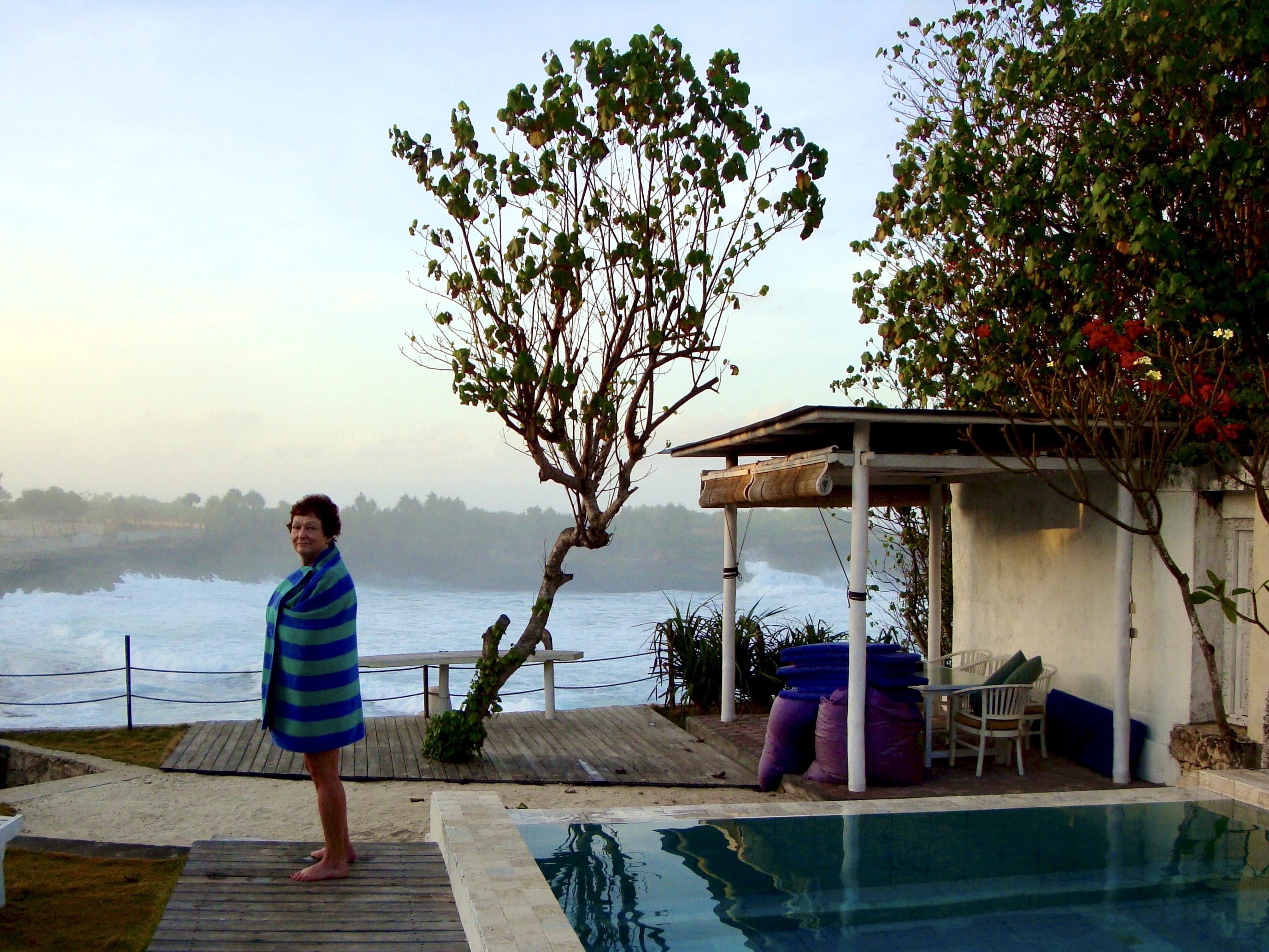 A woman with a towel wrapped around her, standing outside a house with flowing ocean or river water in the background.