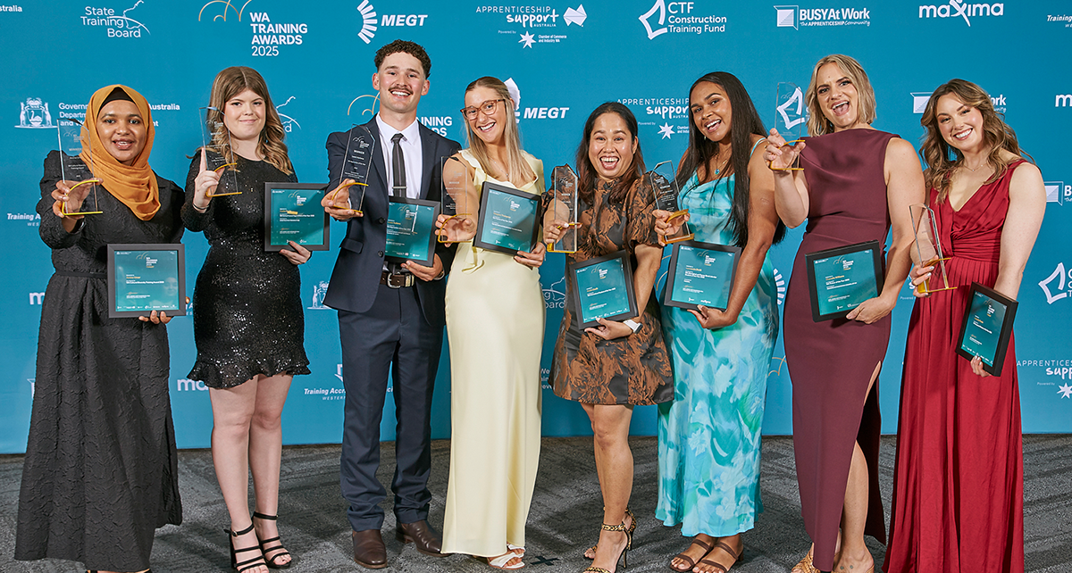 A group of award recipients stand together on a blue event backdrop, each holding a trophy and framed certificate at the WA Training Awards.