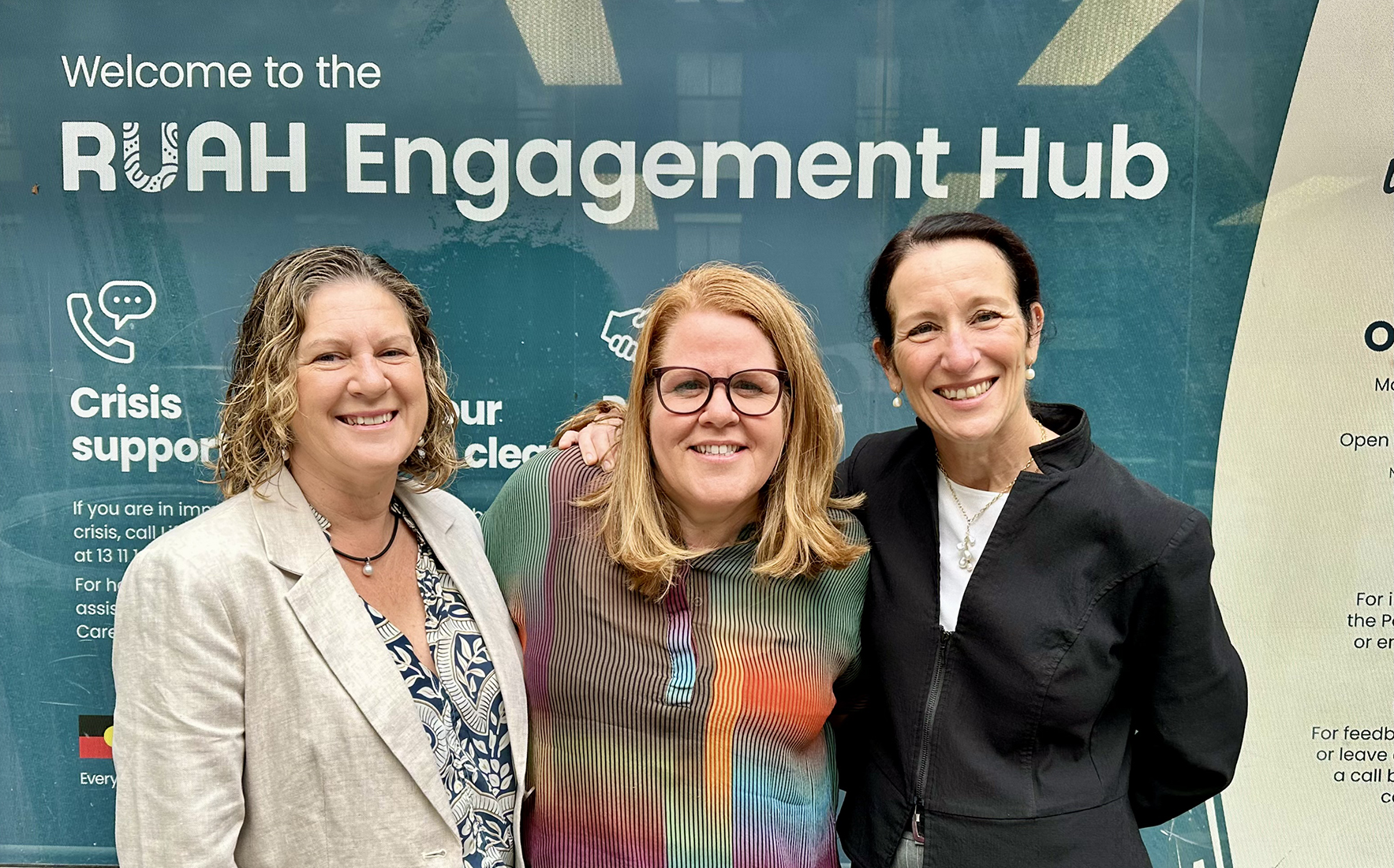 Three women smiling to camera with a banner behind them that reads "Welcome to the RUAH Engagement Hub"