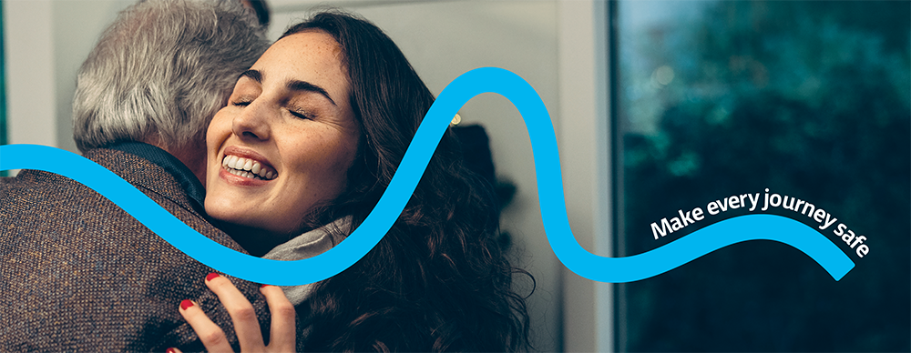 A man and a woman hug at the door of their house. A blue line representing the road runs across the page, with the words Make every journey safe.