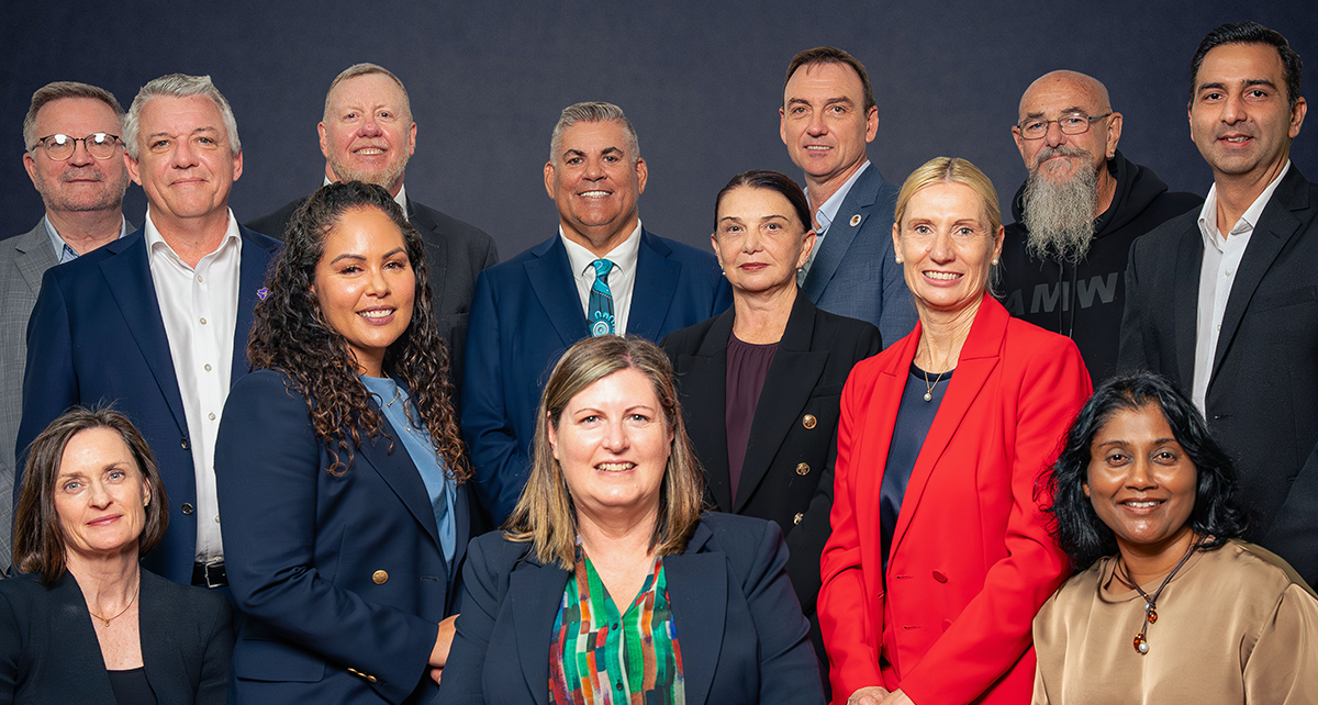 Group portrait of advisory board members standing and seated in two rows against a dark studio background, wearing professional attire and facing the camera.