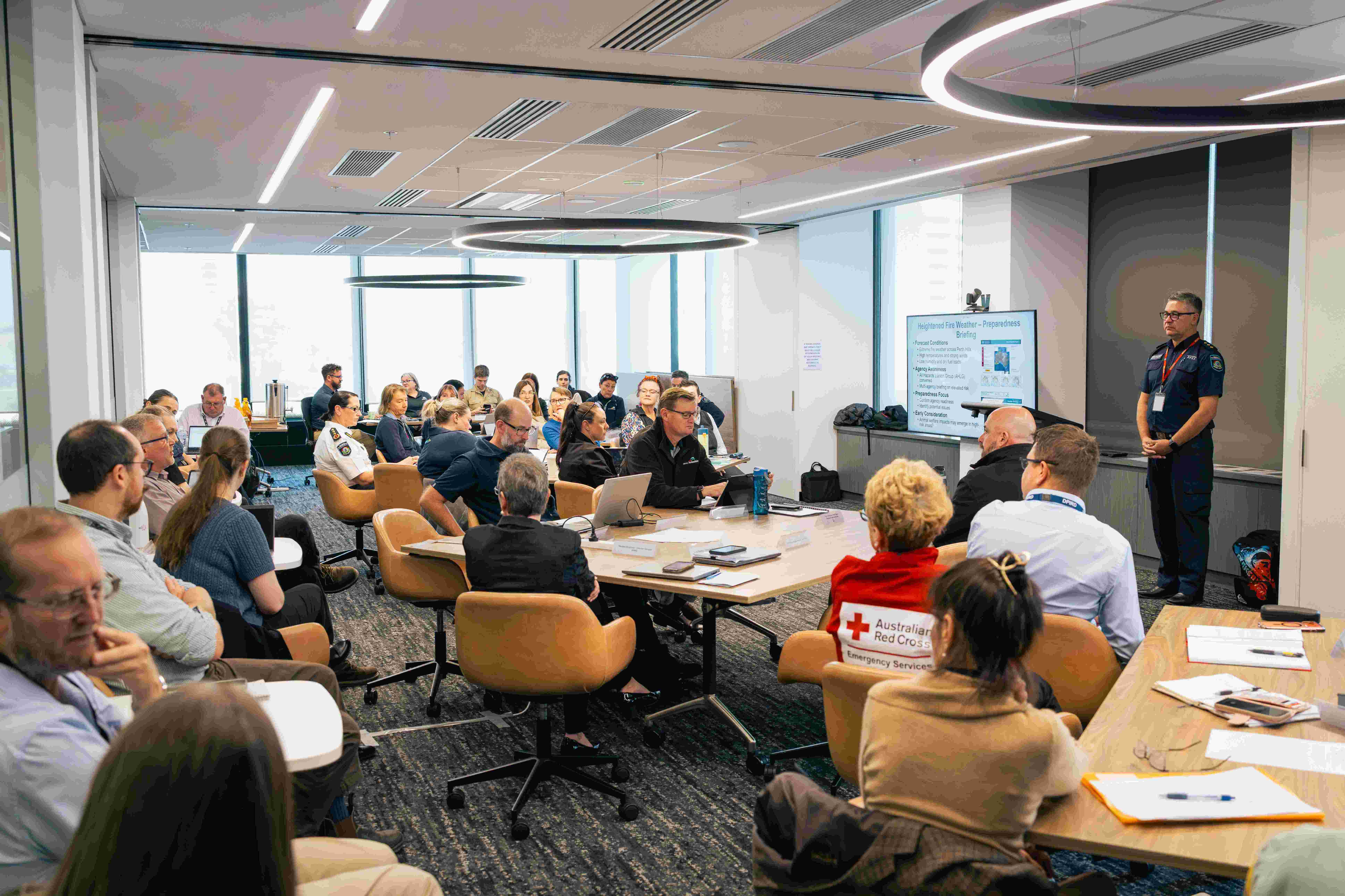 A group of people at tables in a boardroom with a man standing near a whiteboard.