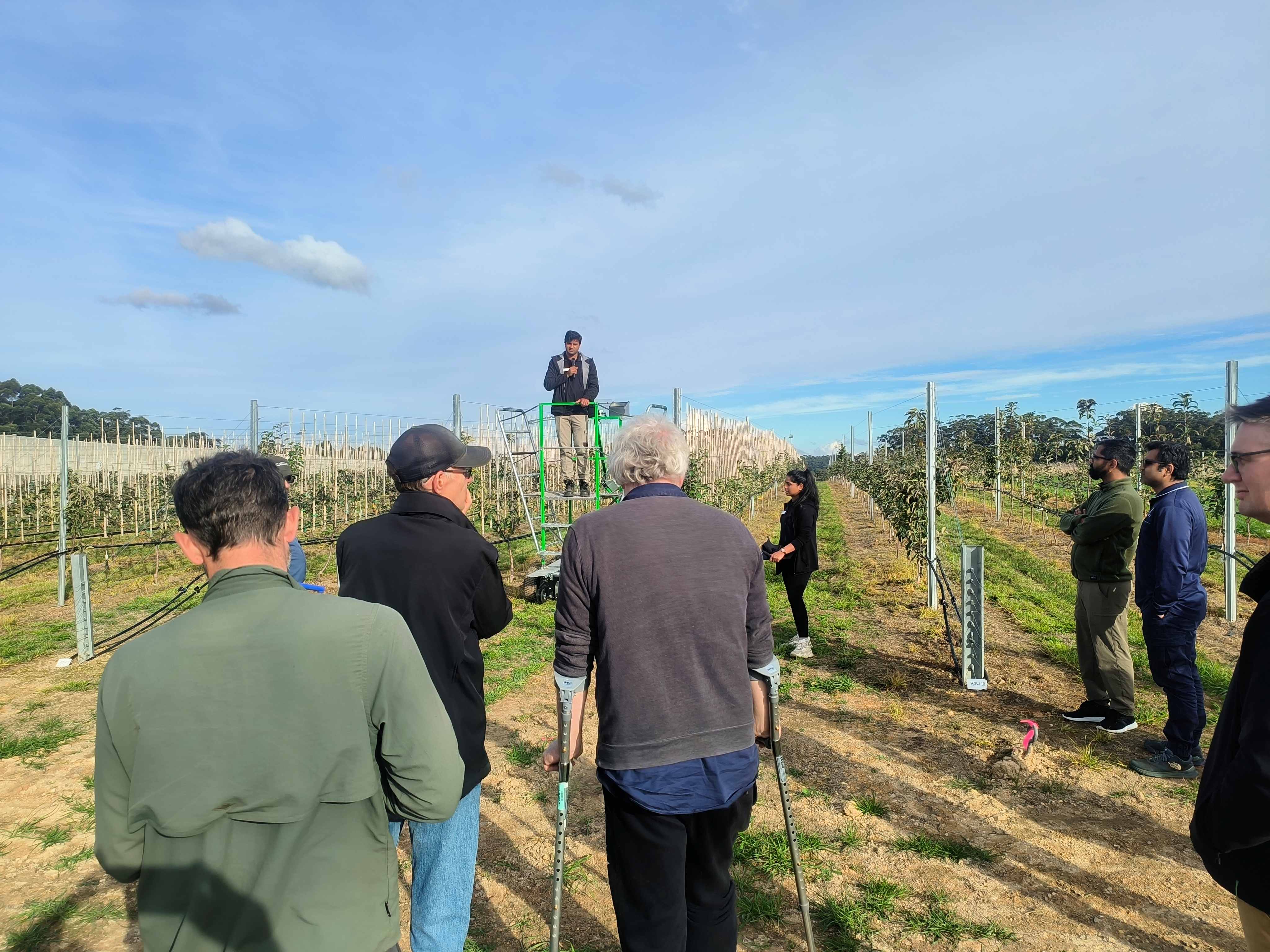 A man standing on front of a platform in an orchard, watched by a crowd.