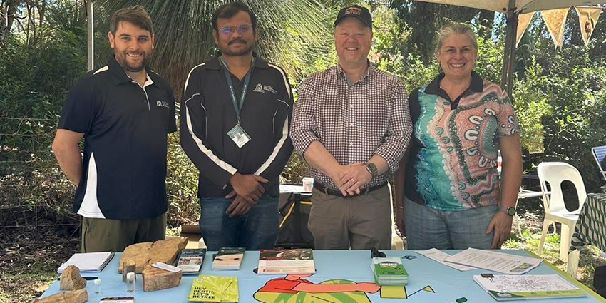 Four people standing behind a Treebate information stall under a gazebo at an outdoor community event, with brochures and displays on the table.