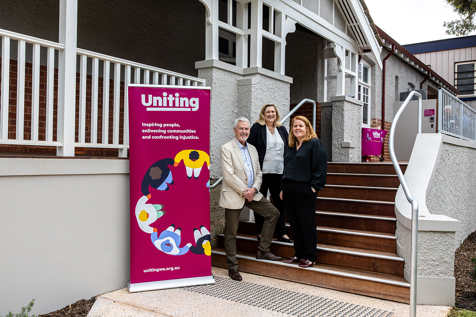 three adult people standing on the front steps of a character home, beside a colourful Uniting WA banner