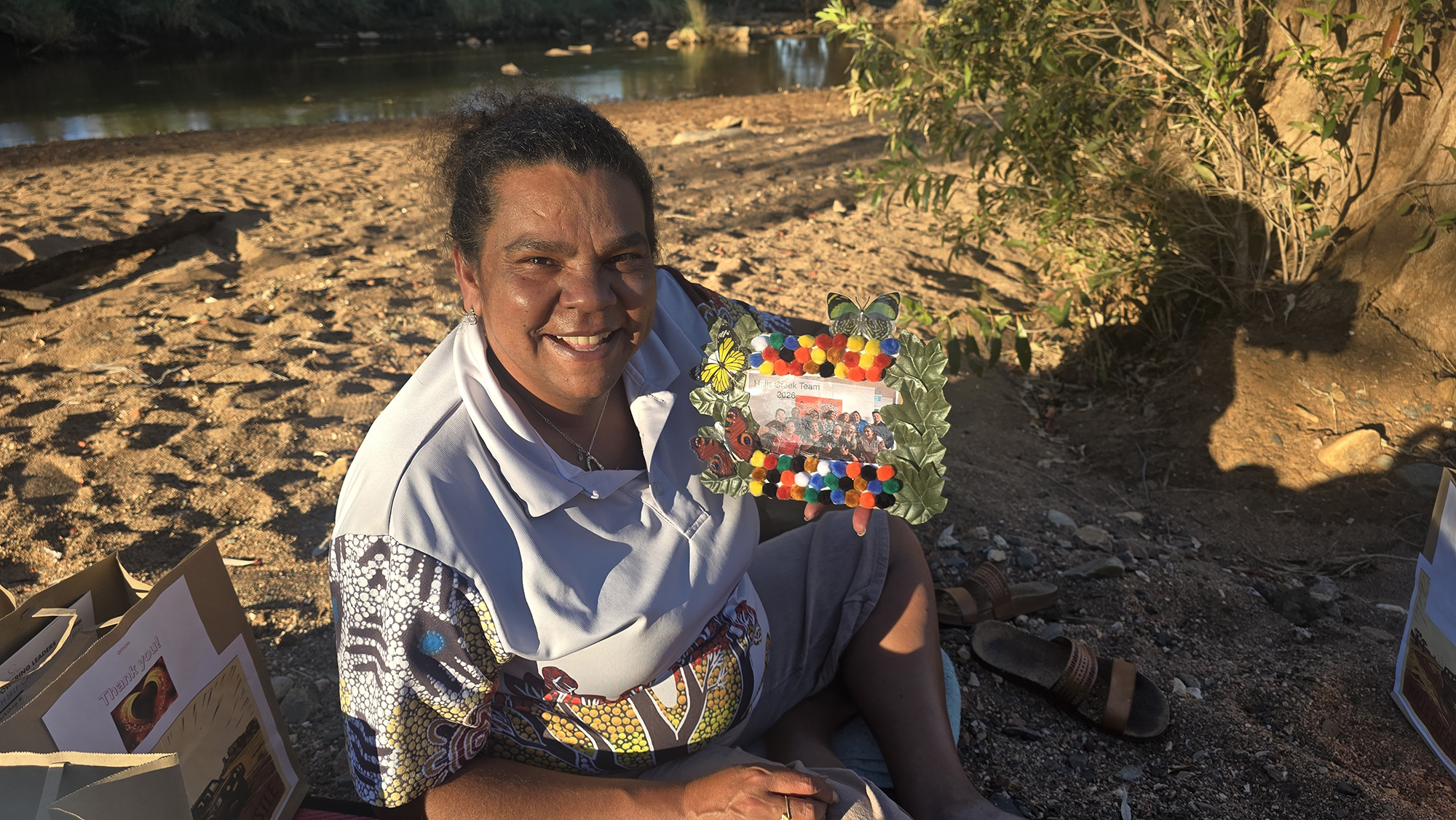 a woman smiling, holding flowers, wearing a polo shirt with Aboriginal artwork. She is sitting on the sand in a natural outback WA environment.
