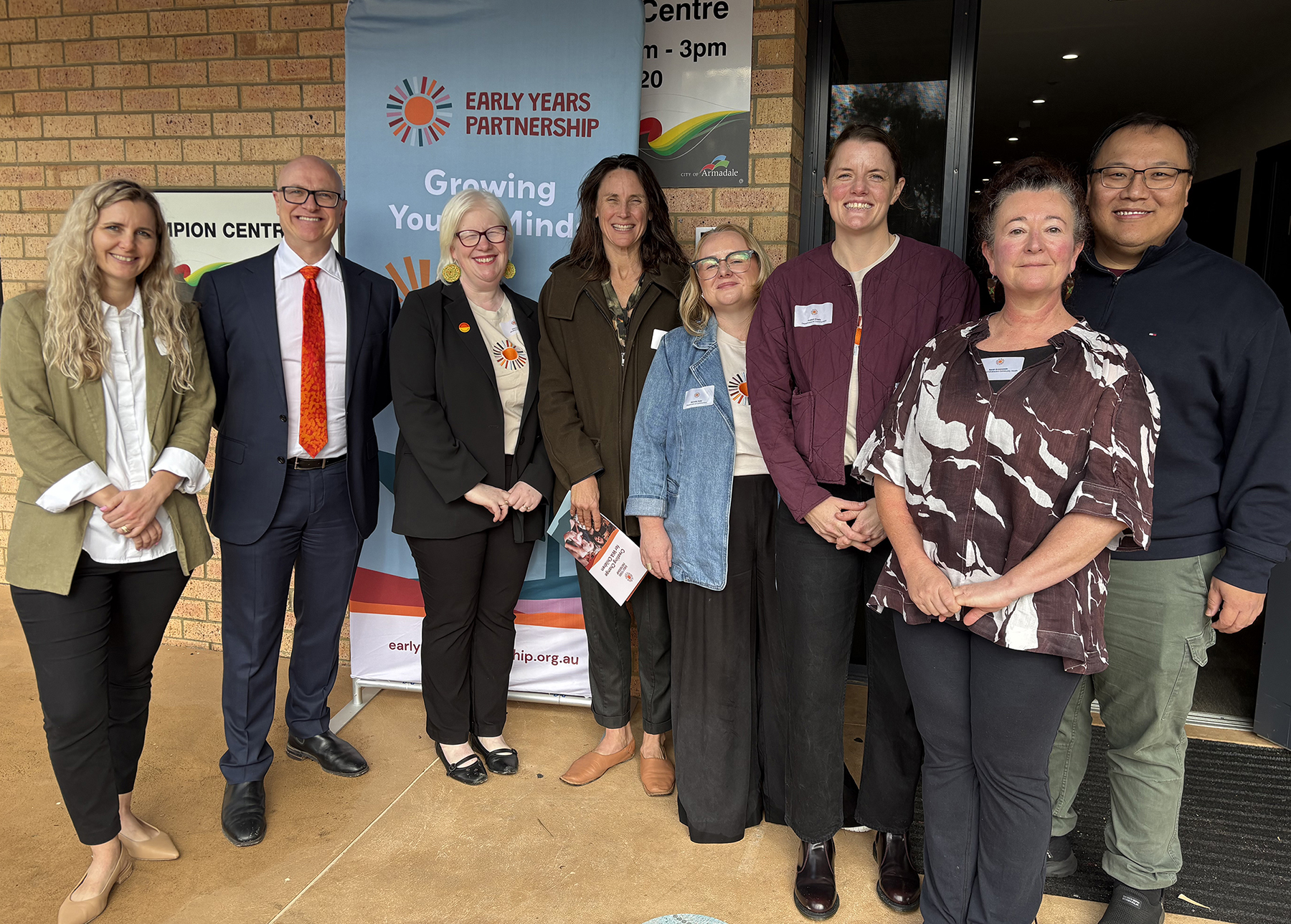 Group shot of seven neatly dressed adults in an outdoor setting, with an Early Years Partnership banner in the background