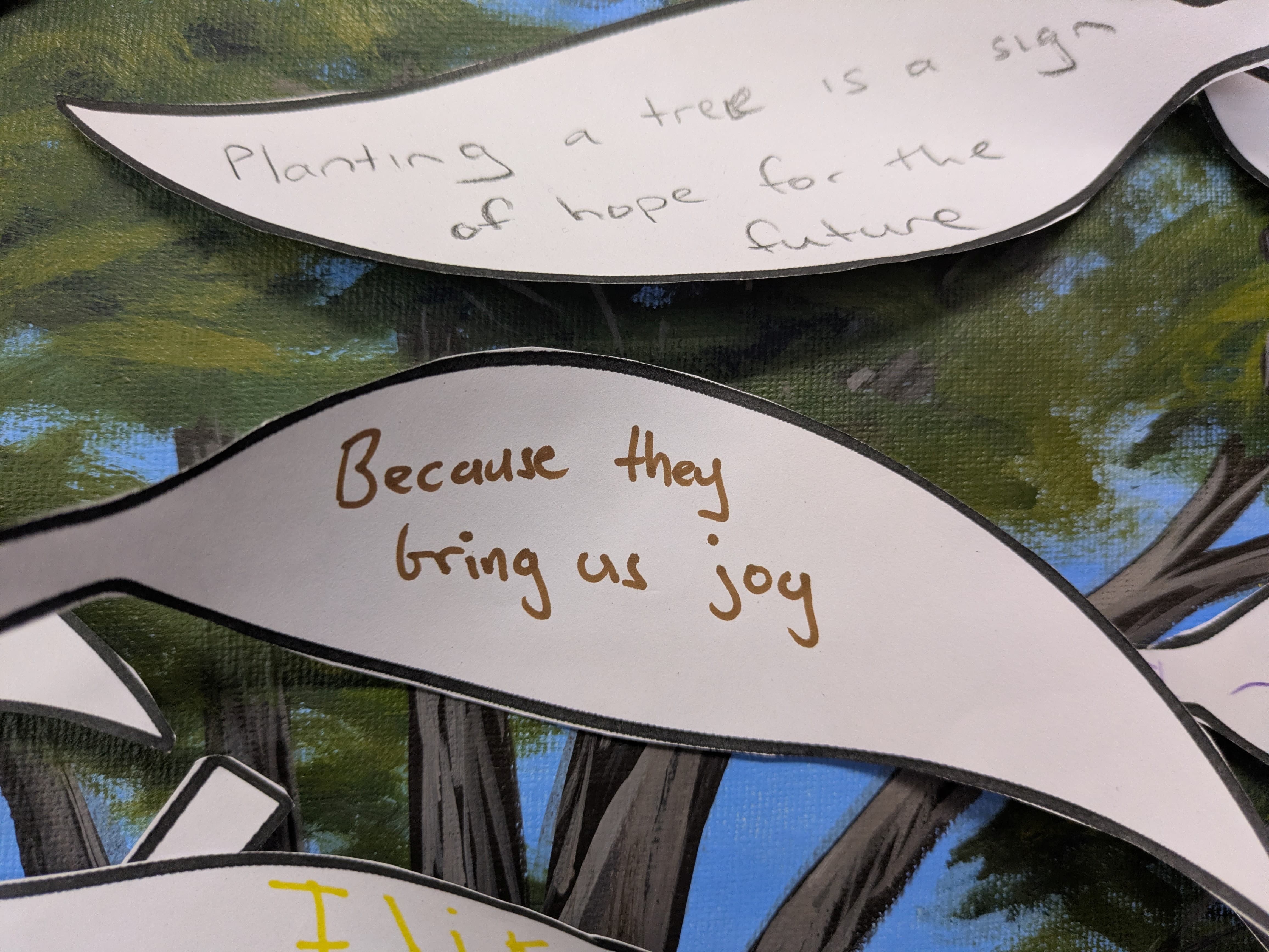 Close-up of paper leaf cut-outs attached to a tree display board. One leaf reads, "Because they bring us joy," and another says, "Plant
