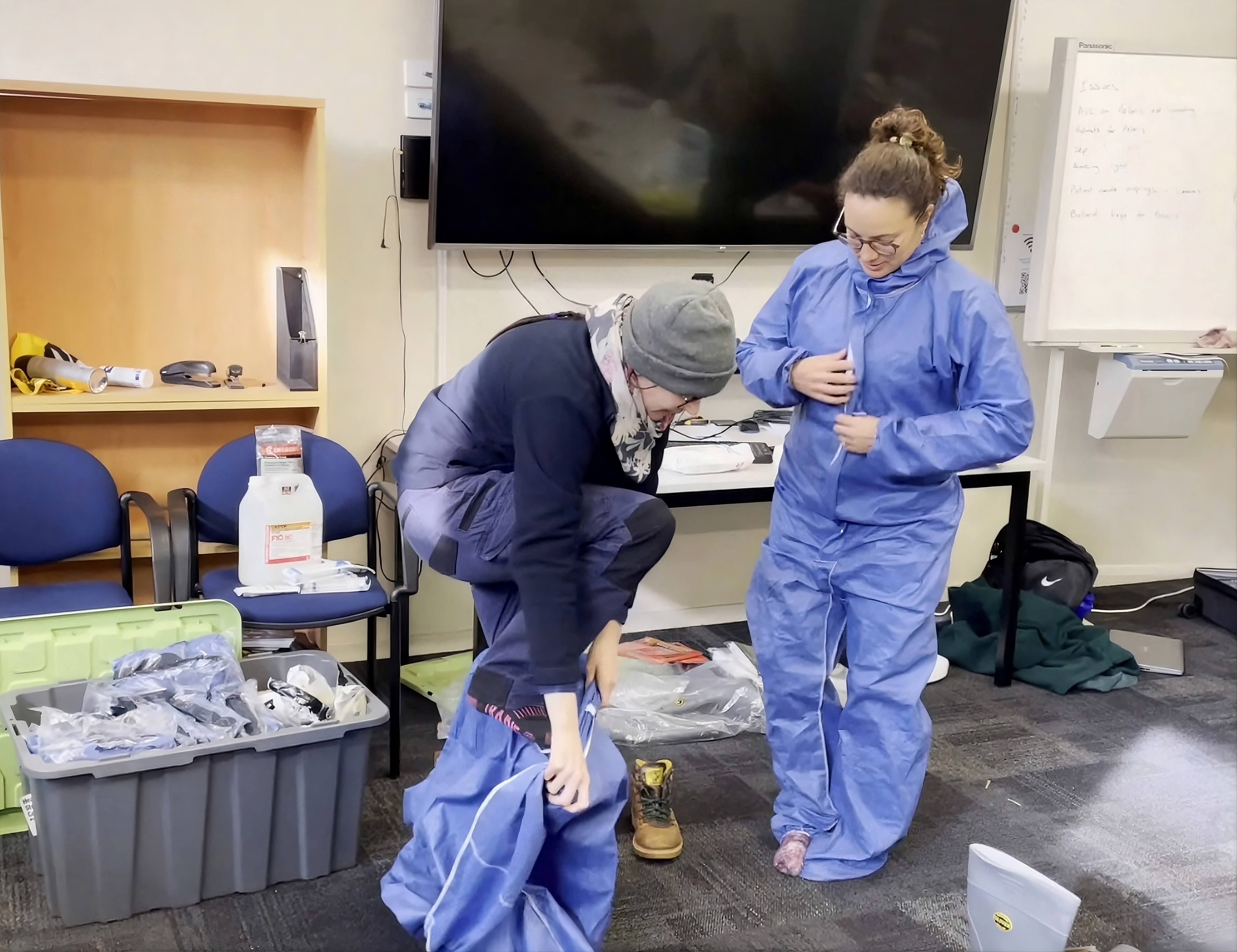 Two women putting on PPE overalls. 