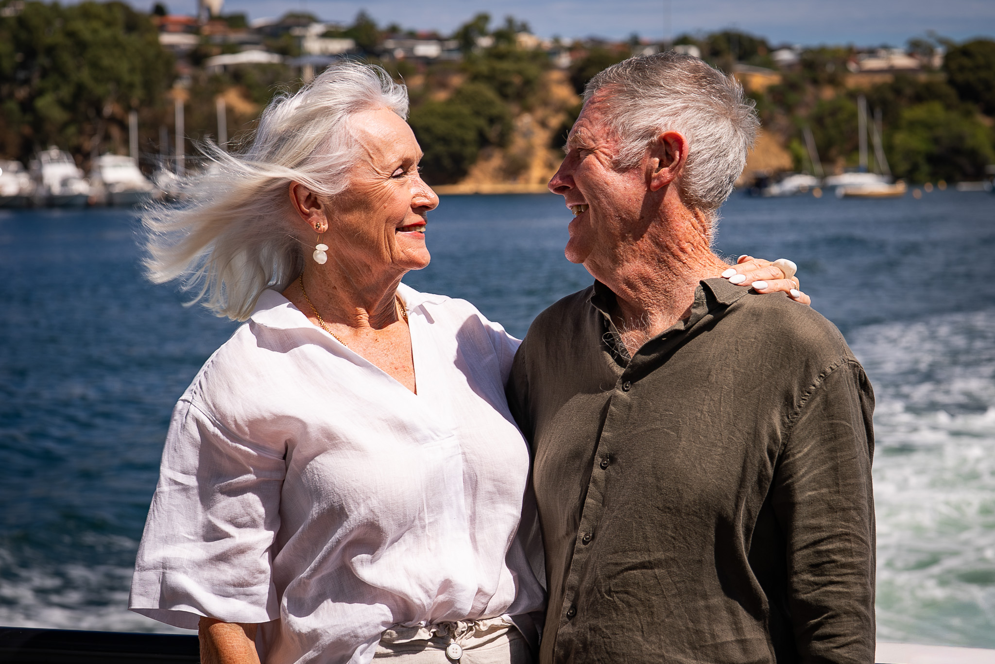 An older man and woman smiling, hugging, with the ocean in the background